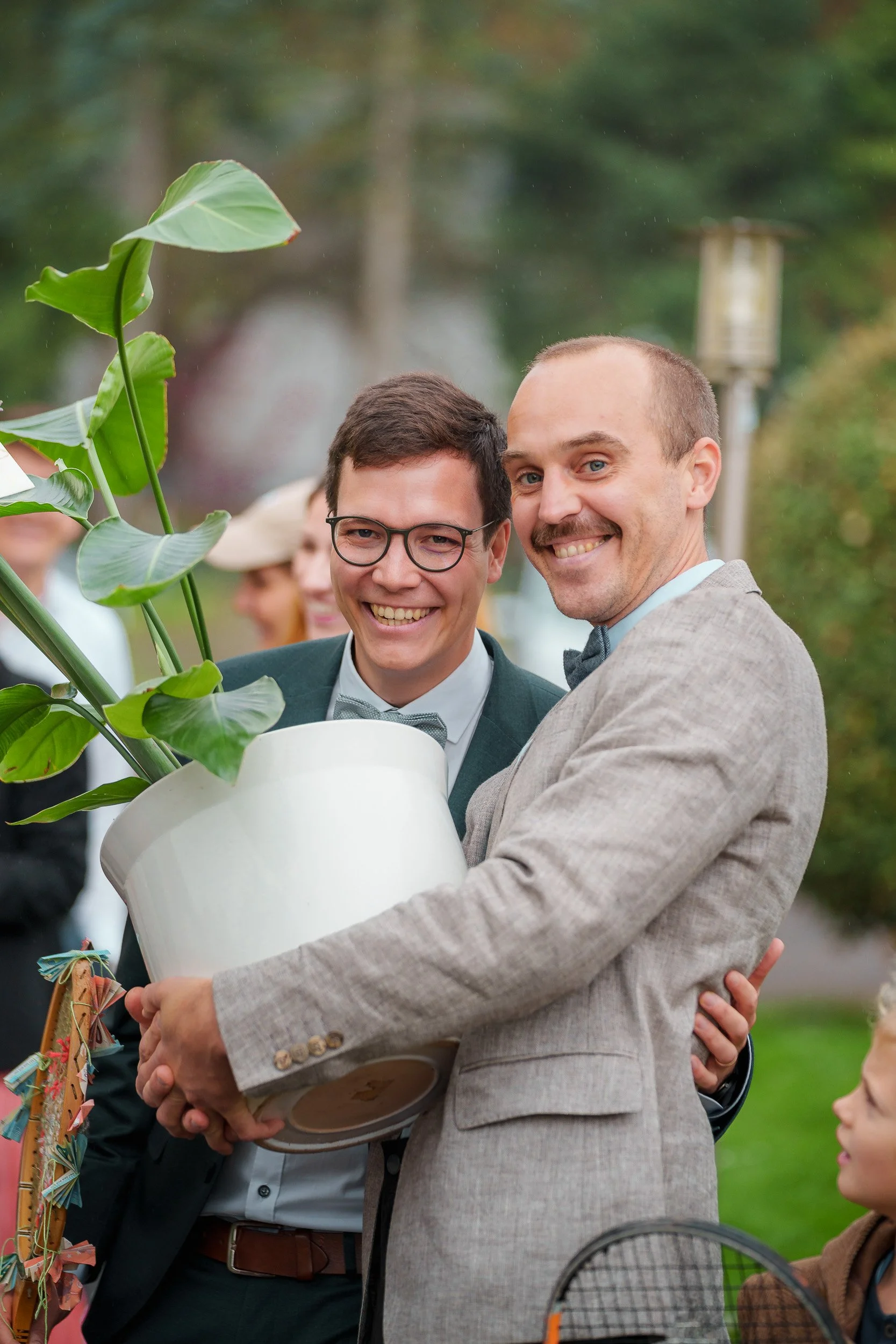 Zwei Männer bei einer Feier halten zusammen einen großen Topf mit einer Pflanze, im Hintergrund weitere Personen und ein Garten.