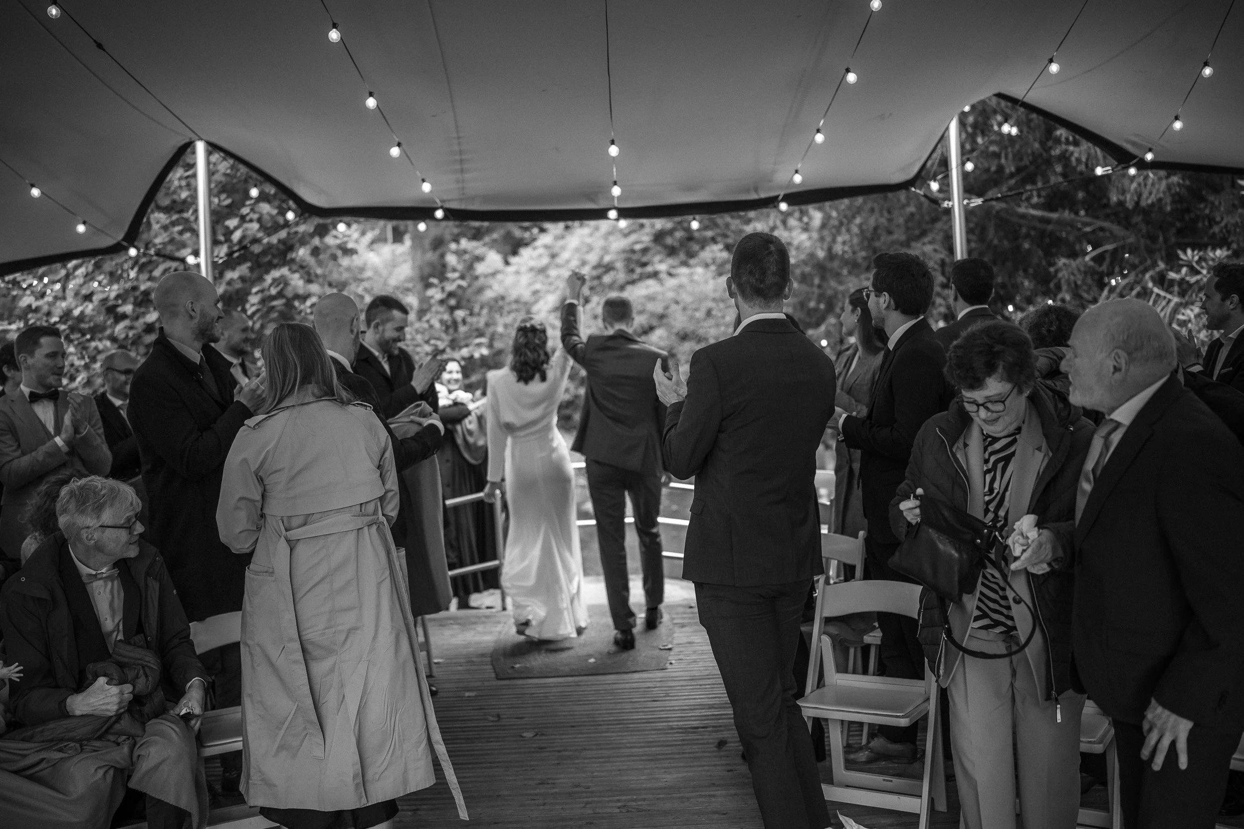 Schwarz-weiß-Fotografie einer Hochzeitsfeier unter einem großen Zelt. Das Brautpaar läuft in die Mitte, umgeben von Gästen, die sich freuen und applaudieren.