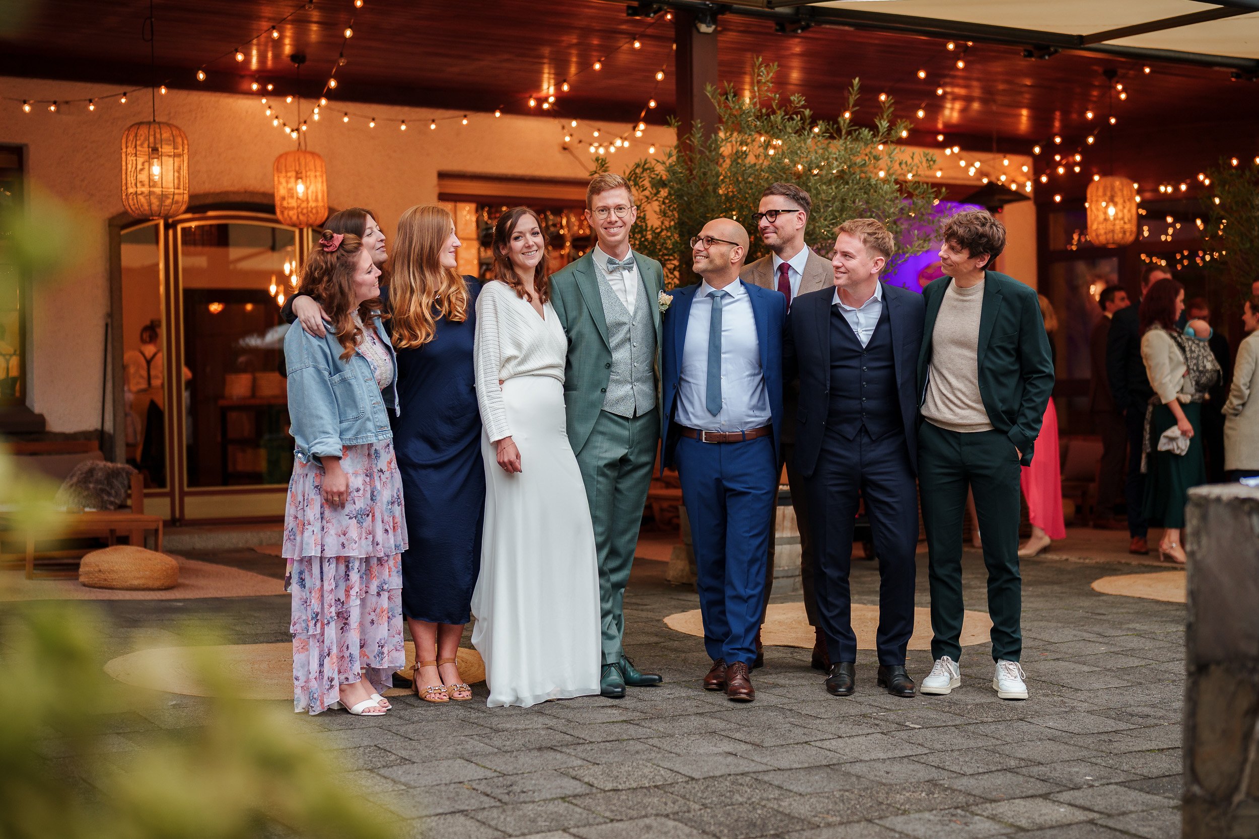 Gruppe von Menschen in formeller Kleidung bei einer Feier, vermutlich Hochzeit, unter Lichterketten im Freien.