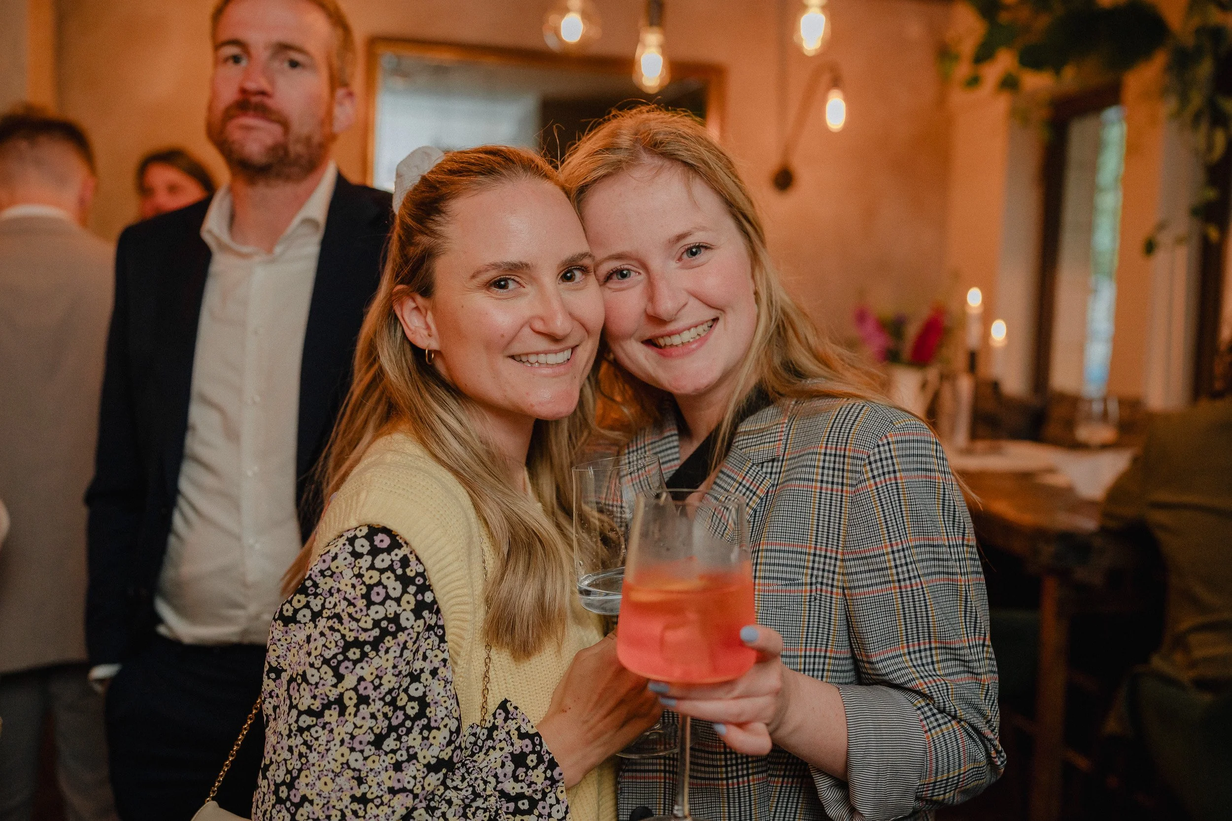 Zwei lachende Frauen bei einer Party, eine hält ein Glas mit rosafarbenem Getränk, im Hintergrund weitere Gäste in einem gemütlichen Raum mit warmem Licht.
