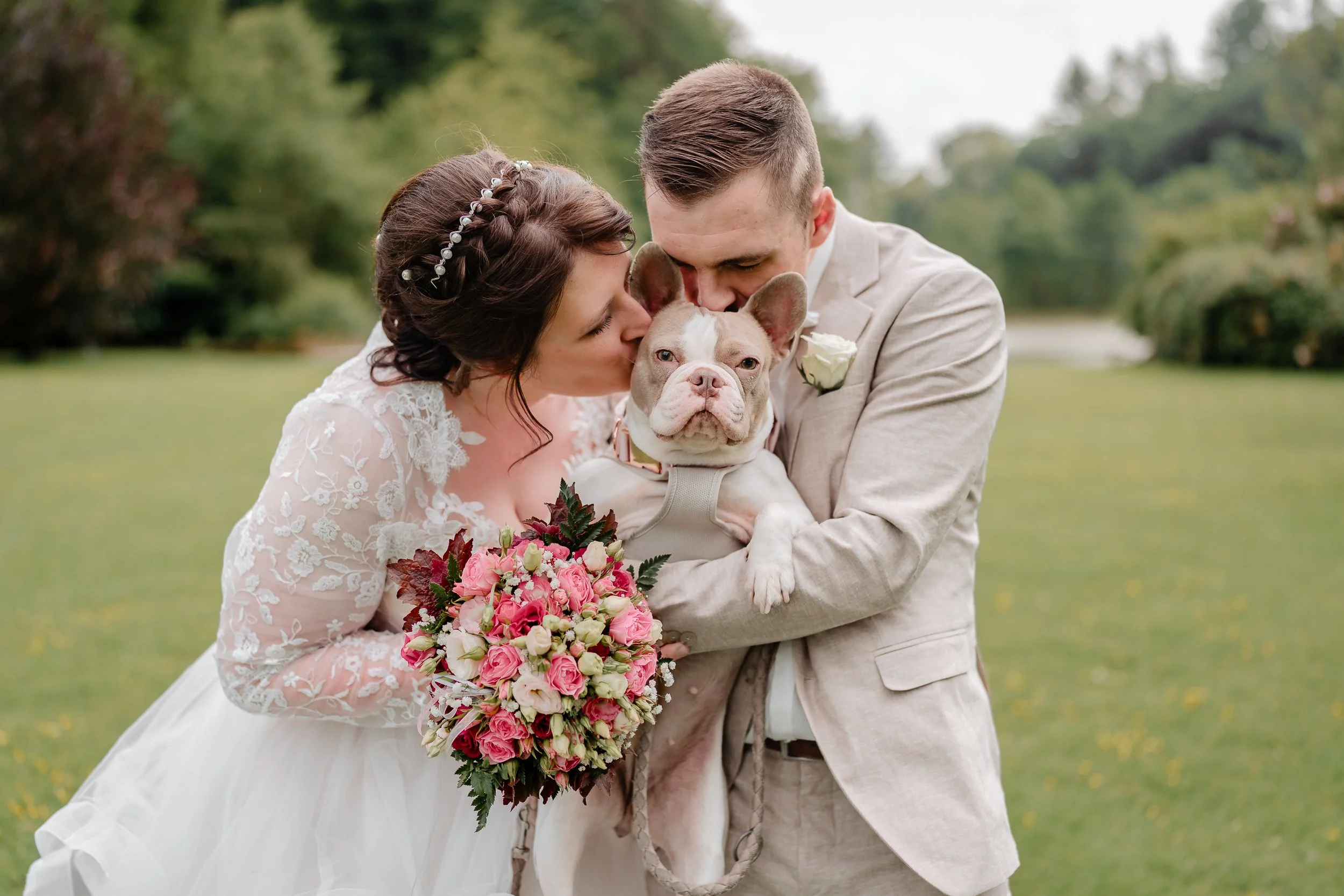 Ein Brautpaar umarmt und küsst einen französischen Bulldoggen-Hund bei einer Hochzeit im Freien, die Braut hält einen Blumenstrauß, der Bräutigam trägt einen weißen Blumenanstecker.