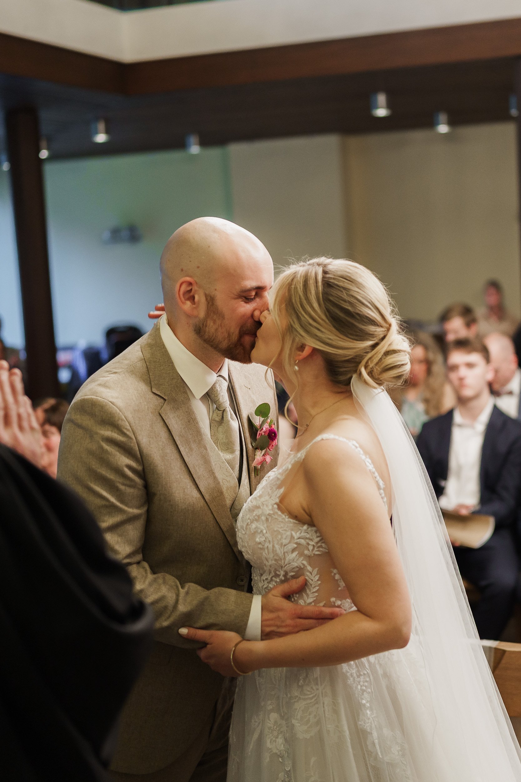 Ein Brautpaar bei der Hochzeit, sie küssen sich im Gottesdienst, im Hintergrund sitzen Gäste und Zeugen.