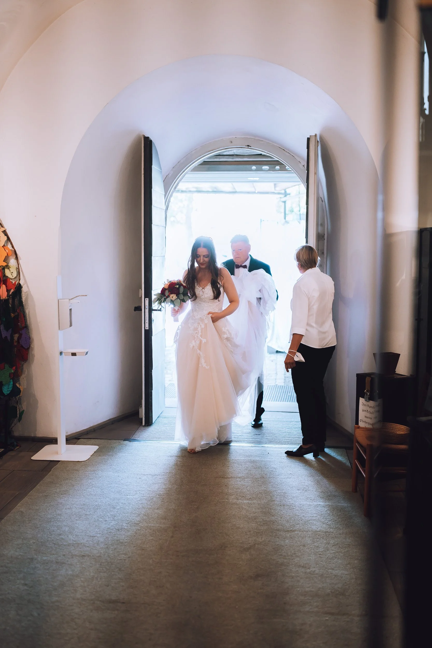 Eine Braut mit Brautkleid, die aus einem Gebäude kommt, gefolgt von einem Bräutigam in Anzug. Zwei andere Personen stehen im Vordergrund, eine davon hat eine weiße Bluse, die andere eine dunkle Hose und Haare.