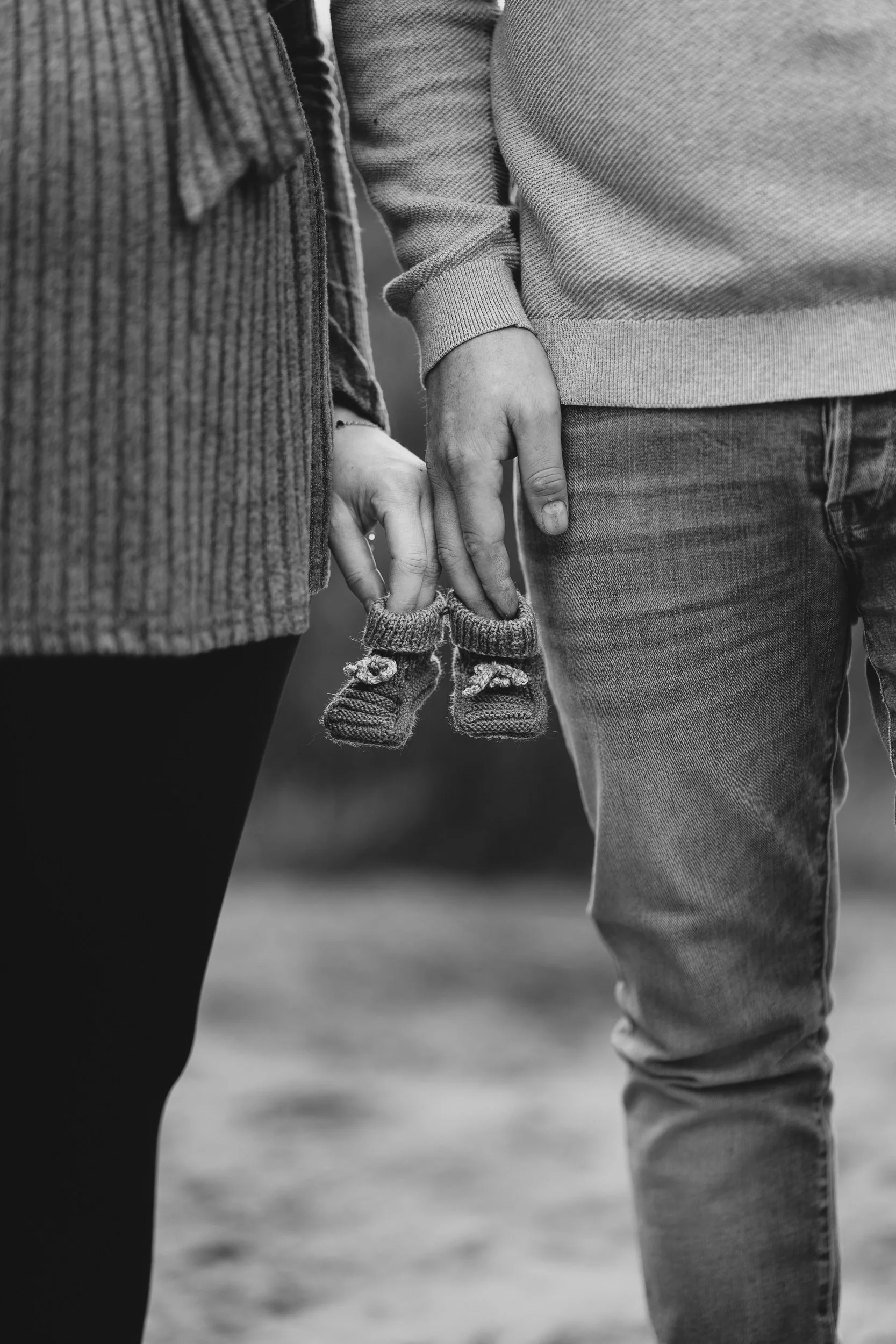 Nahaufnahme eines Paares, das sich an den Händen hält, wobei die Hände im Fokus stehen. Das Paar trägt warme Kleidung, und in ihrer Hand hängt ein Paar kleine Babyschühchen.
