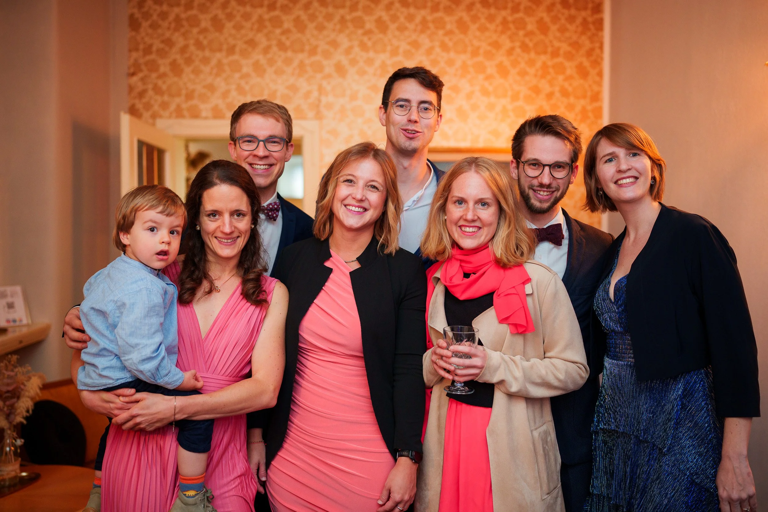 Gruppe von acht Menschen, darunter ein Kind, bei einer Feier in einem Wohnzimmer mit goldgelber Wand und offener Tür. Alle lächeln und sind elegant gekleidet.