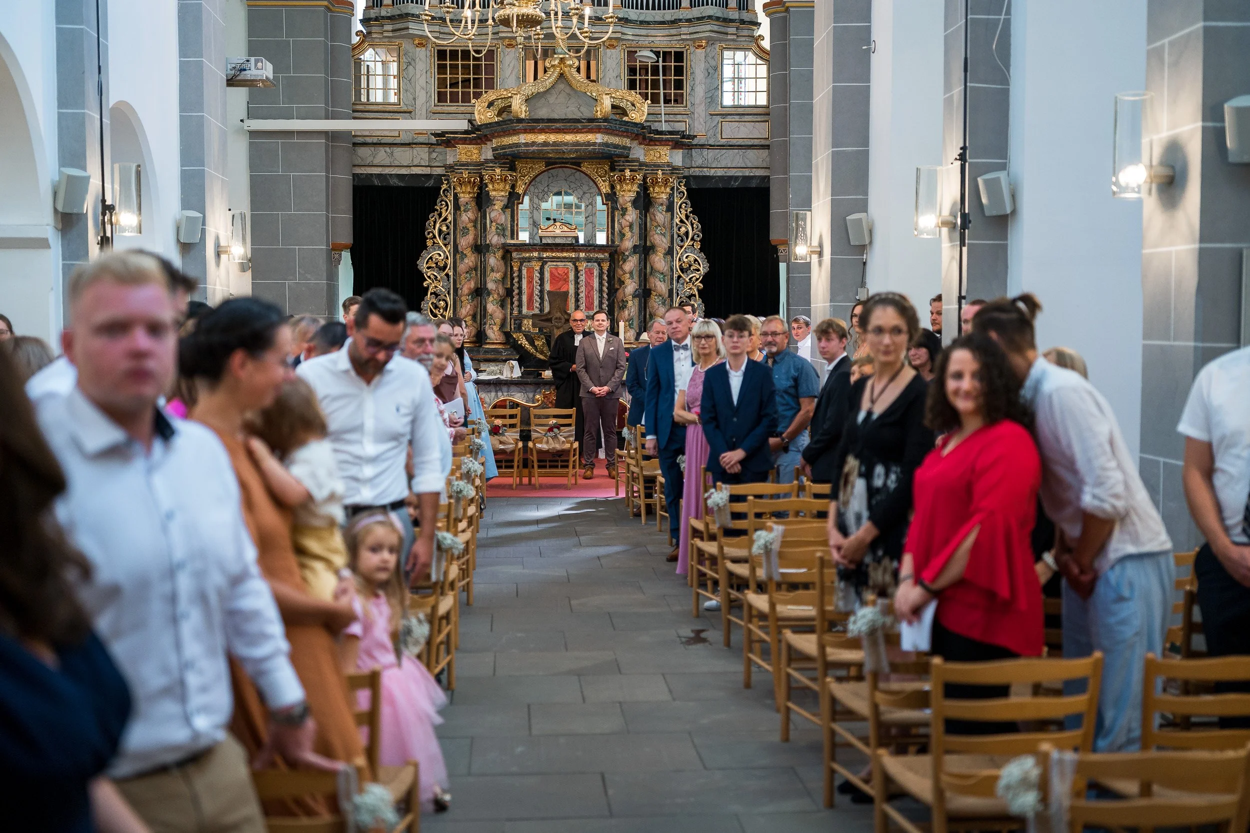 Menschen in einem Festsaal, die wahrscheinlich auf eine Zeremonie warten, mit goldverzierten Barock-Architektur im Hintergrund.