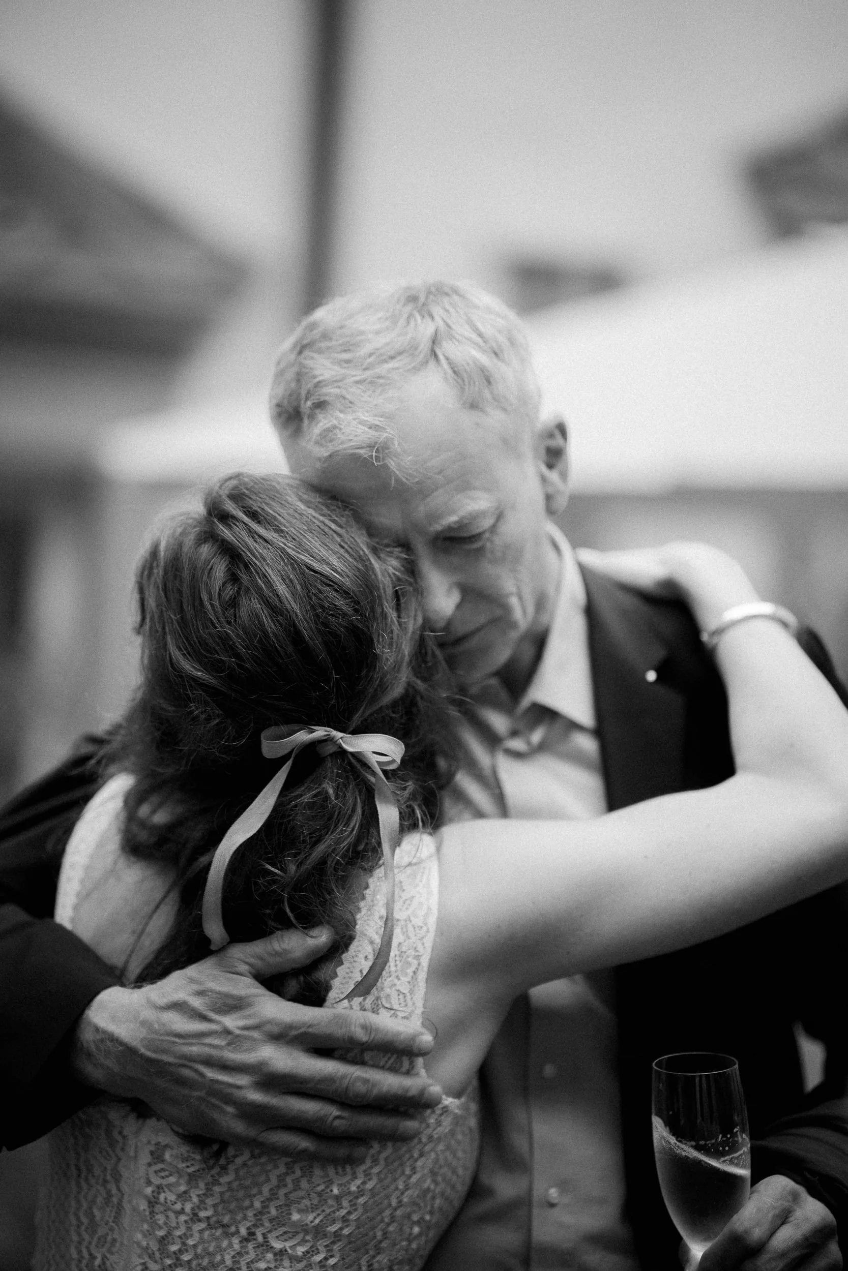 Schwarz-weiß-Foto von zwei Menschen, die sich umarmen, eine ältere Person und eine jüngere Person, die junge Frau trägt ein Kleid mit Spitzendesign und eine Schleife im Haar, während die ältere Person einen Anzug trägt.