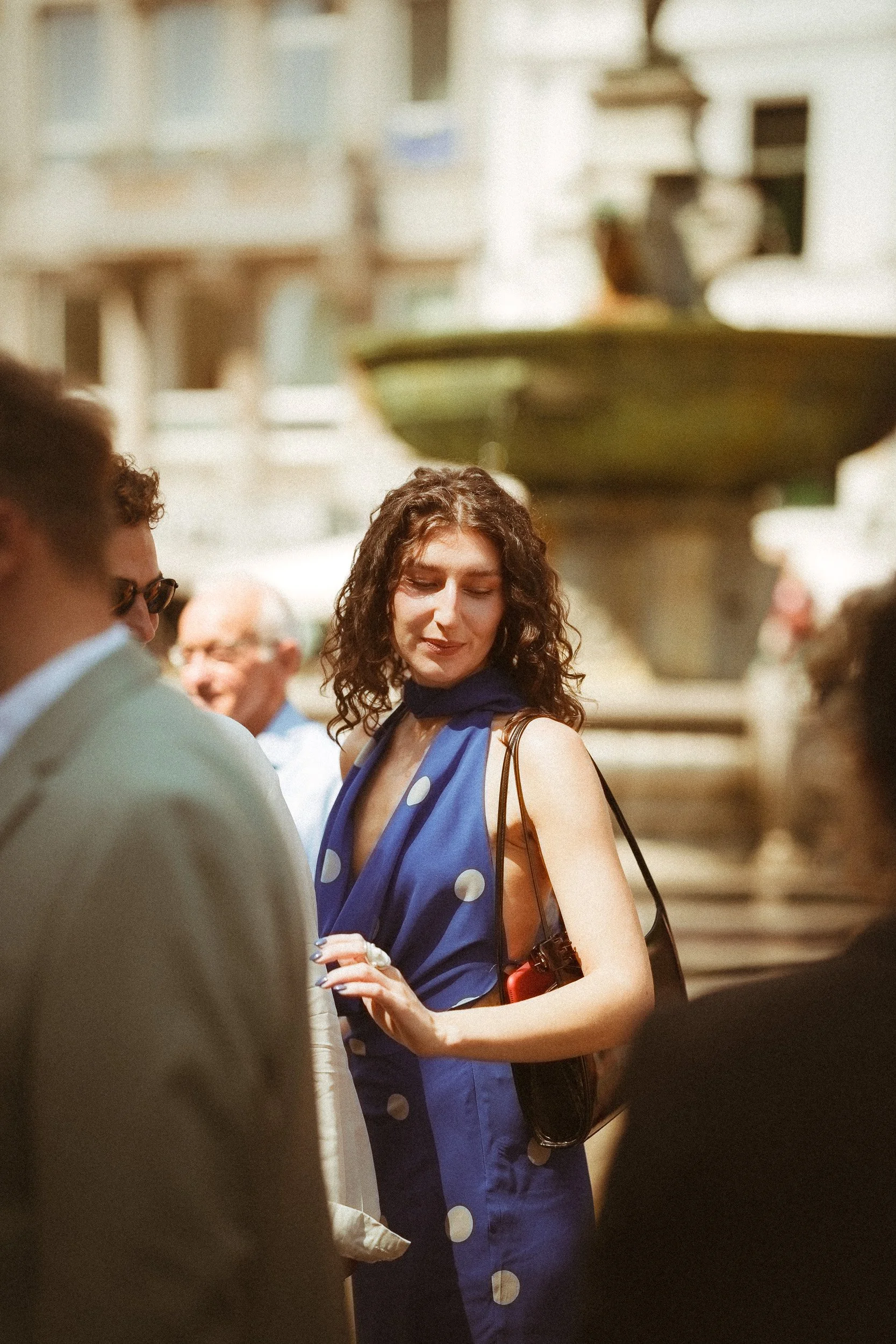 Eine Frau mit lockigem Haar in einem blauen Kleid mit weißen Punkten steht inmitten einer Gruppe von Menschen, im Hintergrund ein Springbrunnen in einer Stadt.