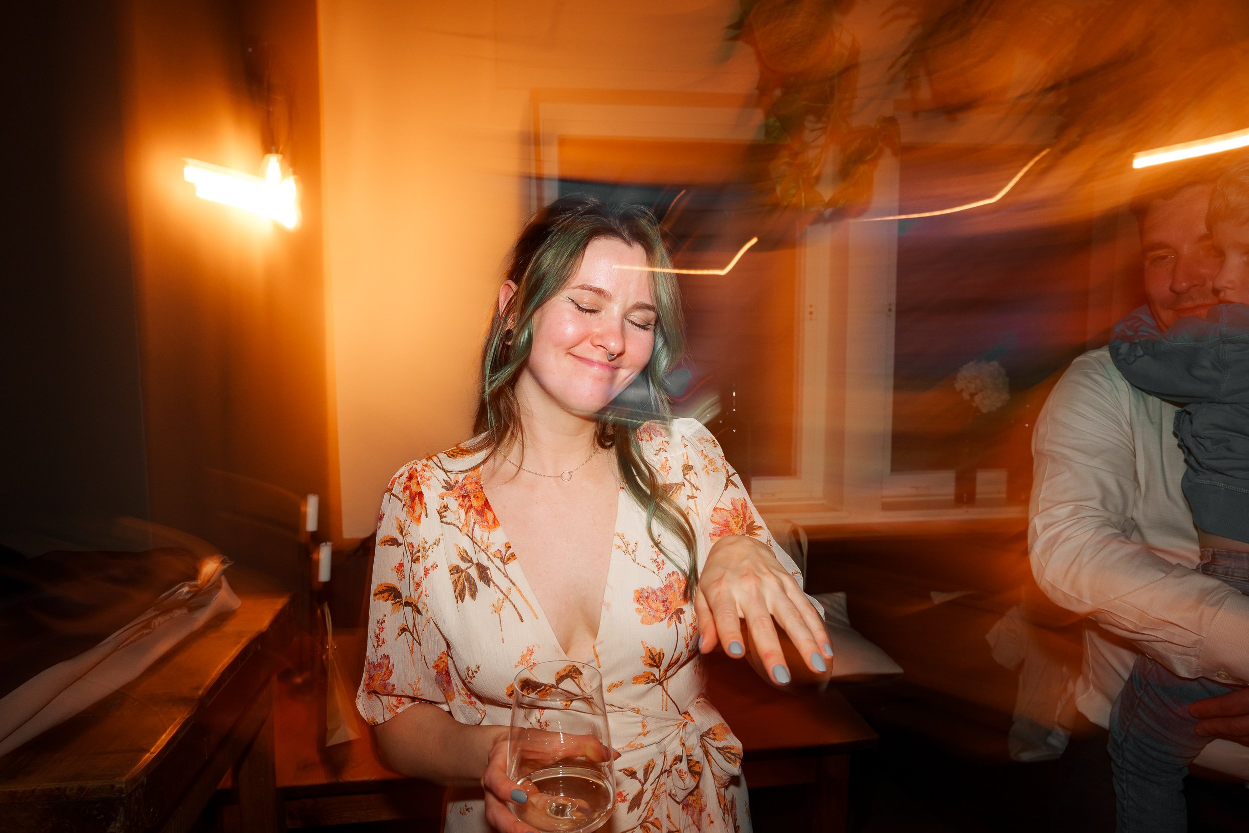 Junge Frau mit langem Haar und Blumenmuster-Kleid lehnt an einem Tisch, hält ein Glas Wein und hat die Augen geschlossen, während sie genießt. Im Hintergrund sind verschwommene Lichter und eine Person, die ein Kind hält.