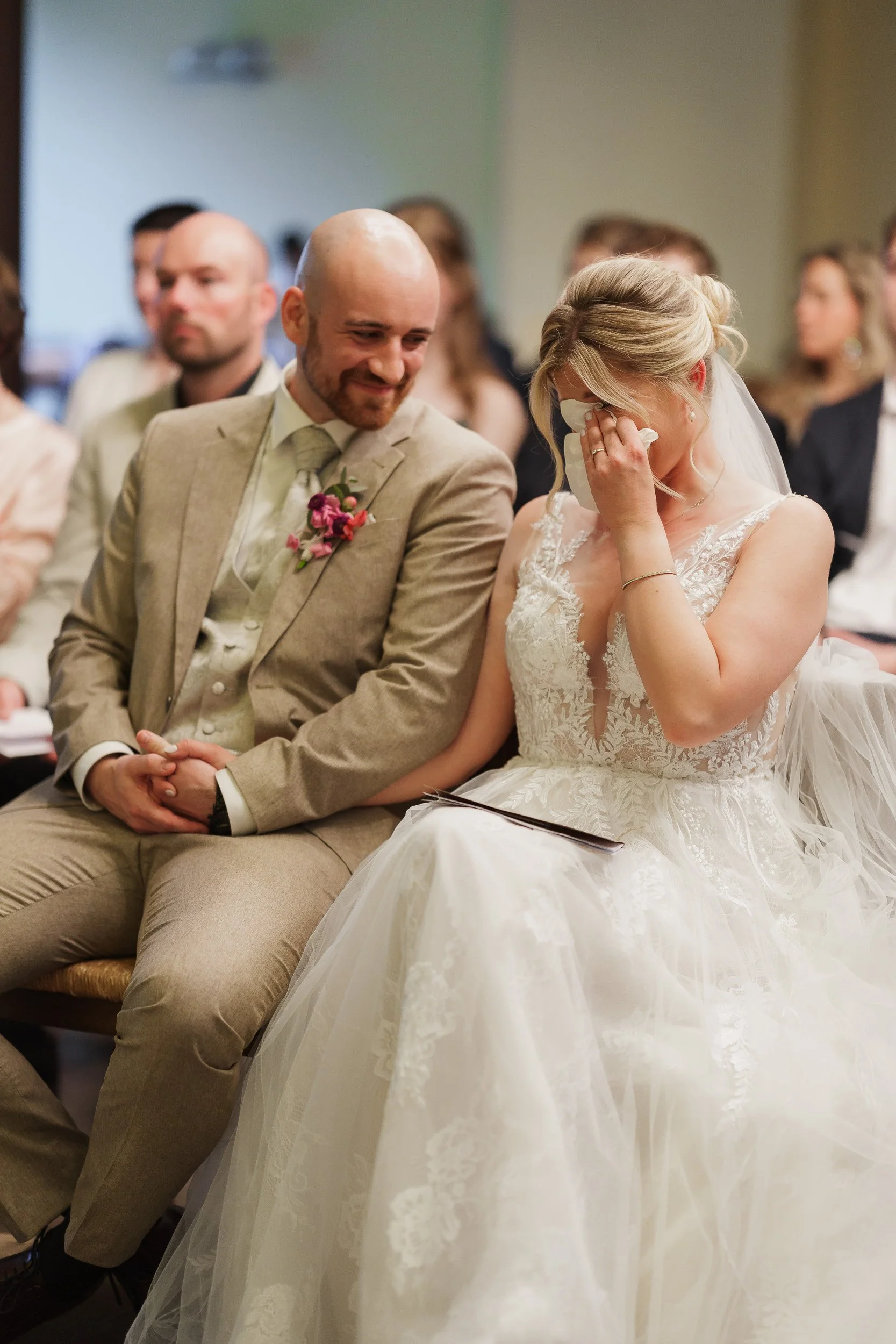 Ein Brautpaar bei ihrer Trauung, die Braut ist emotional und hält ein Taschentuch an ihr Gesicht, der Bräutigam lächelt liebevoll.