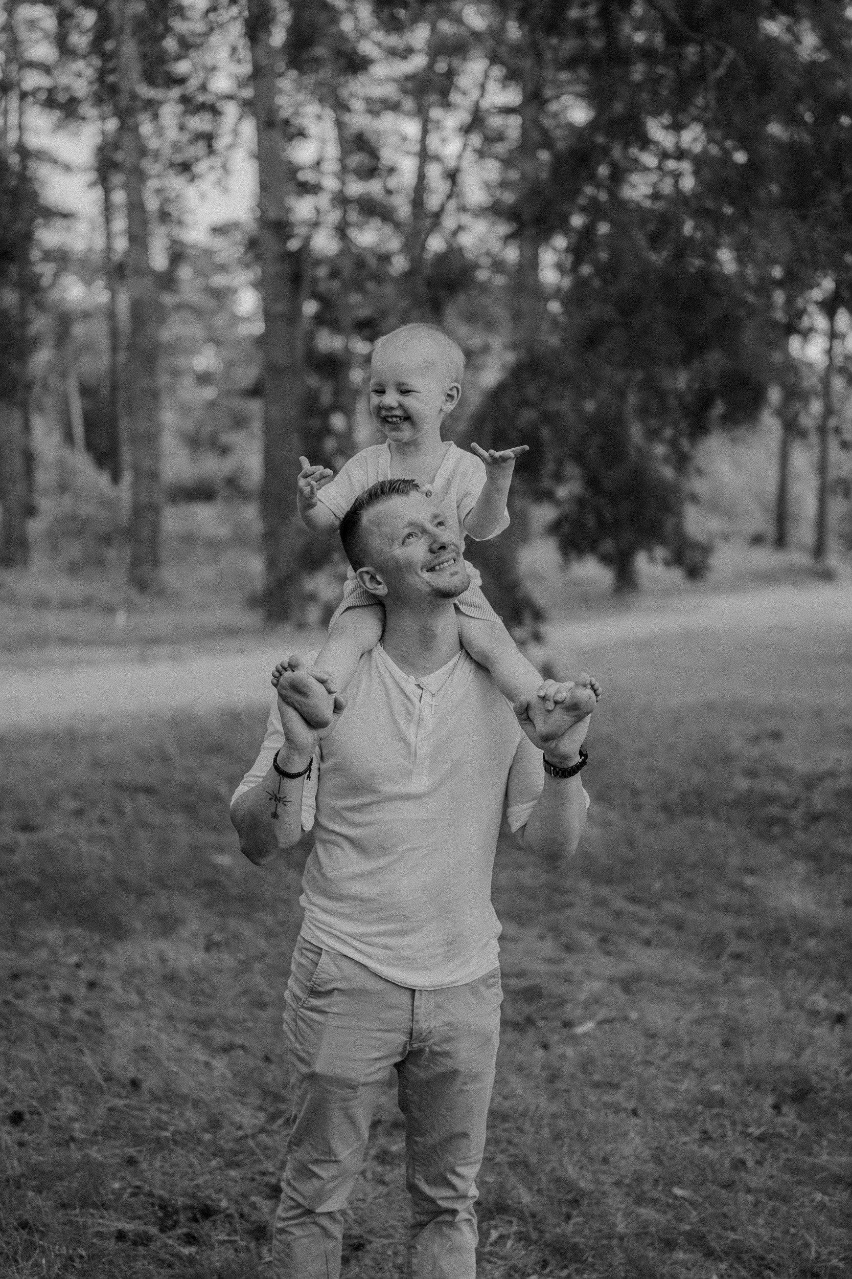 Ein Mann trägt ein Kind auf den Schultern im Freien, umgeben von Bäumen, beide lachen und haben Spaß.