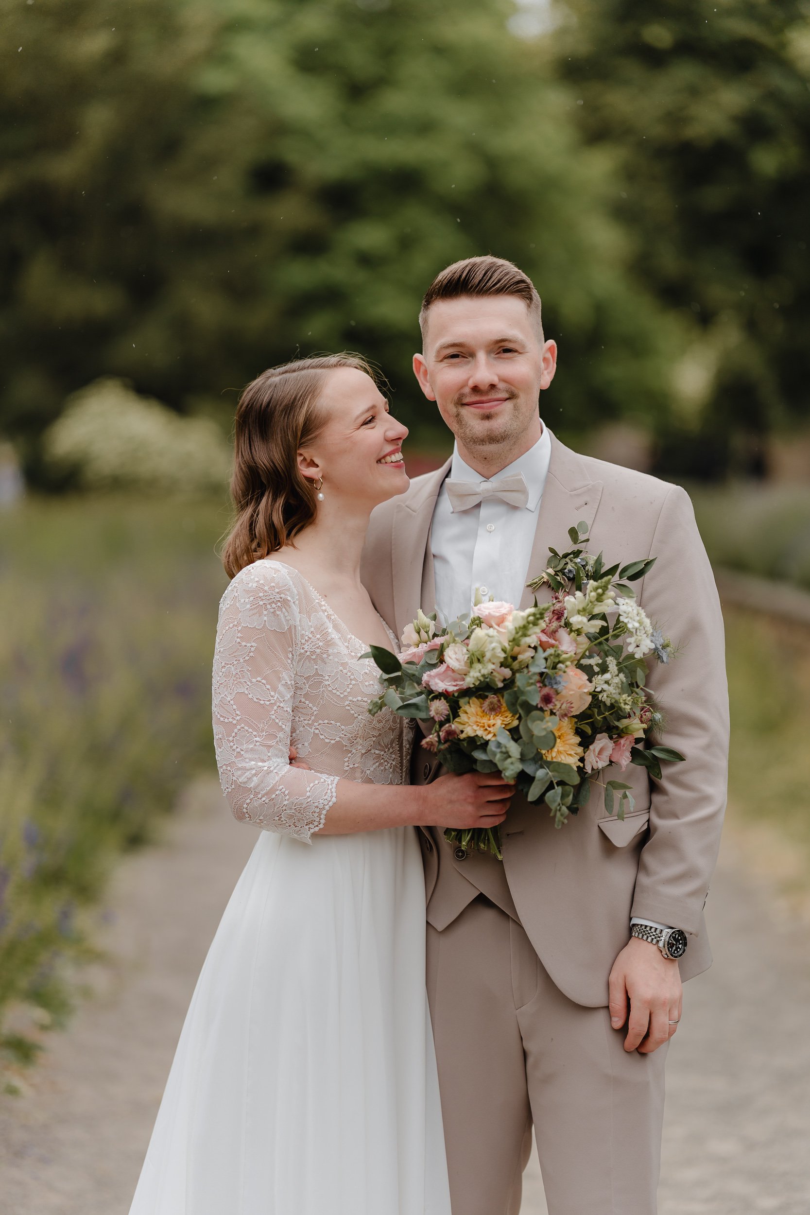 Ein Brautpaar steht im Freien, die Braut hält einen Blumenstrauß, die Braut trägt ein Kleid mit Spitzenärmeln, der Bräutigam trägt einen Anzug mit Fliege, sie lächeln sich an.