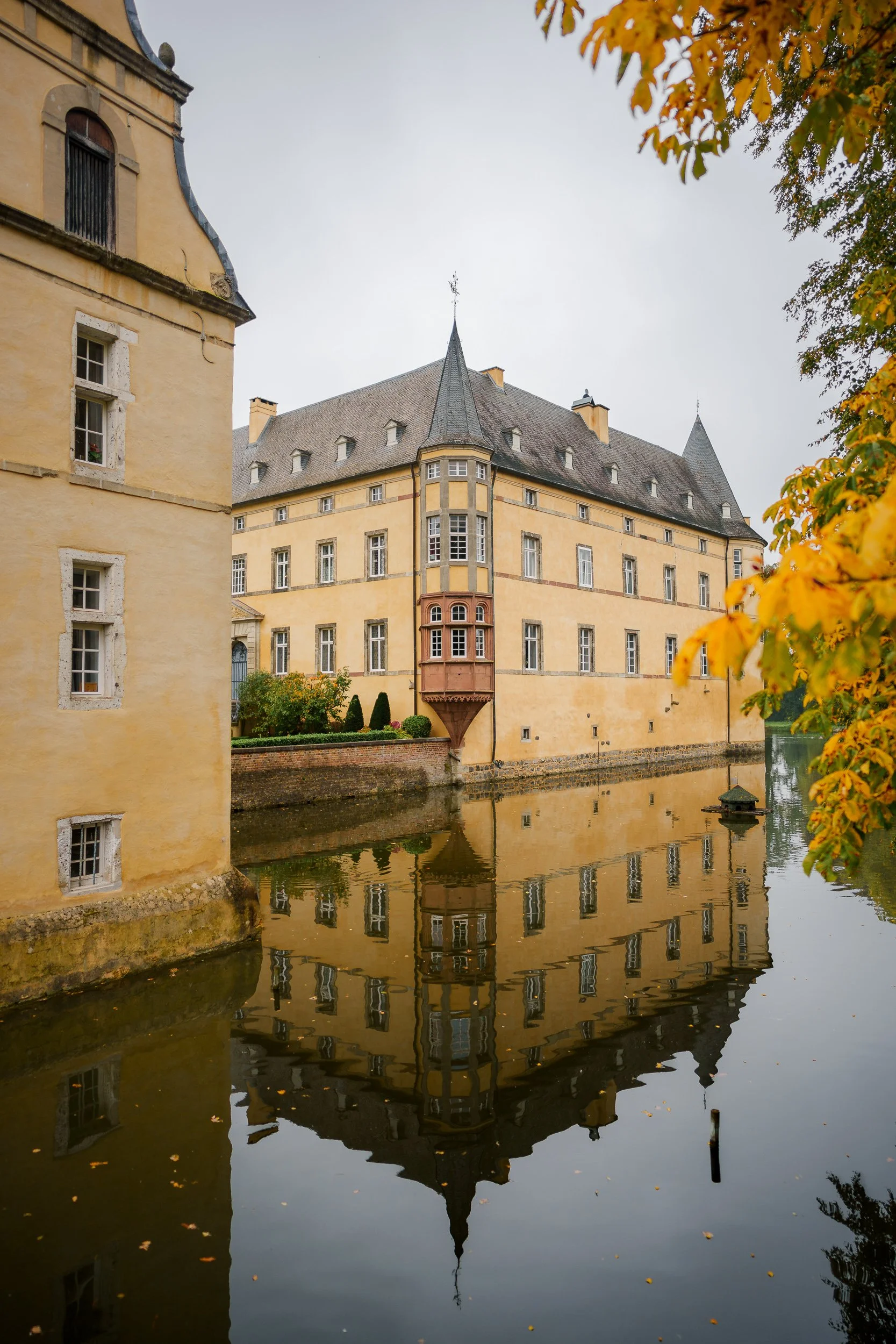Ein historisches Schloss mit gelber Fassade und spitzen Türmen, das sich im Wasser spiegelt, umgeben von Herbstblättern.