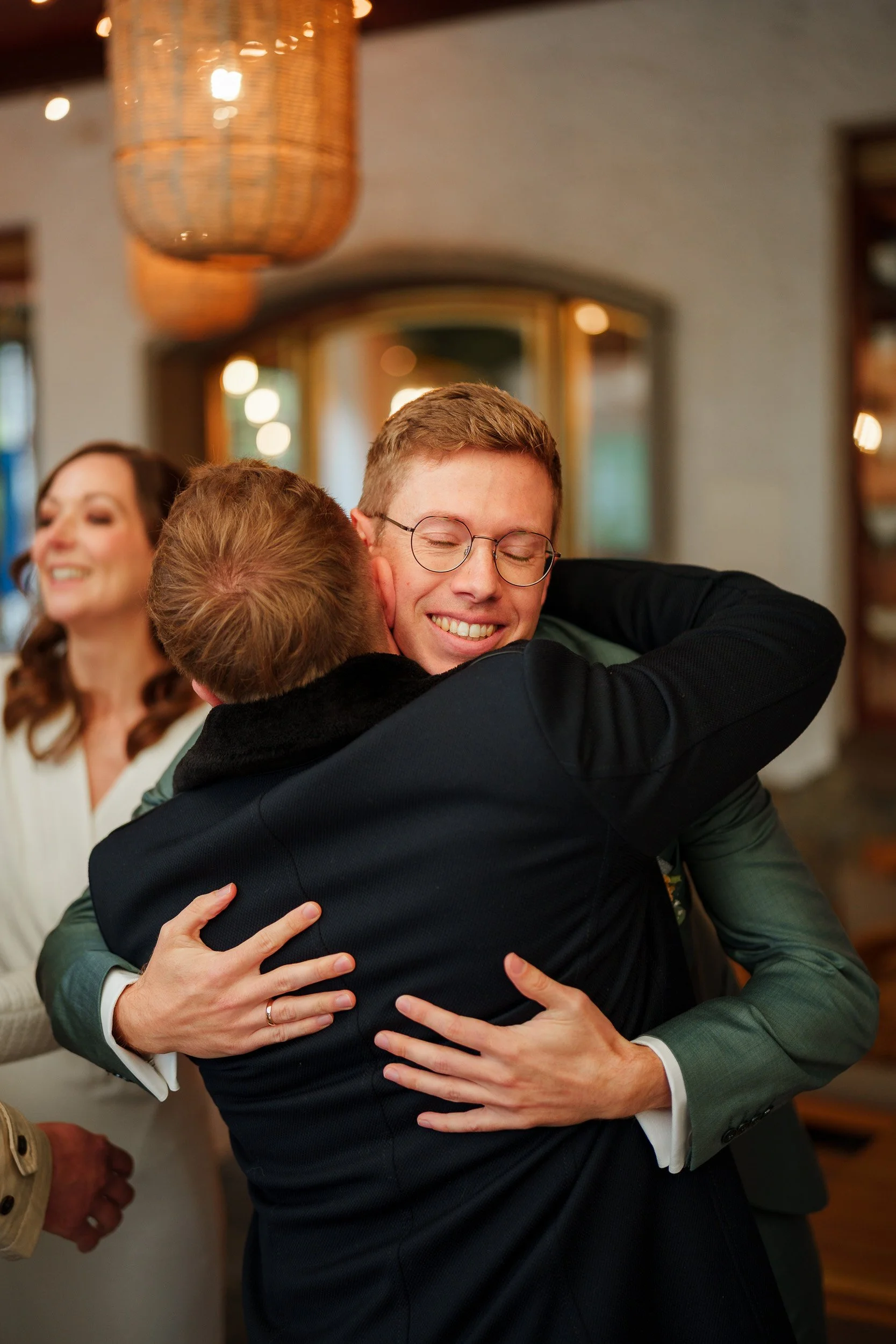 Zwei Männer umarmen sich freudig bei einer Feier, im Hintergrund eine lachende Frau.