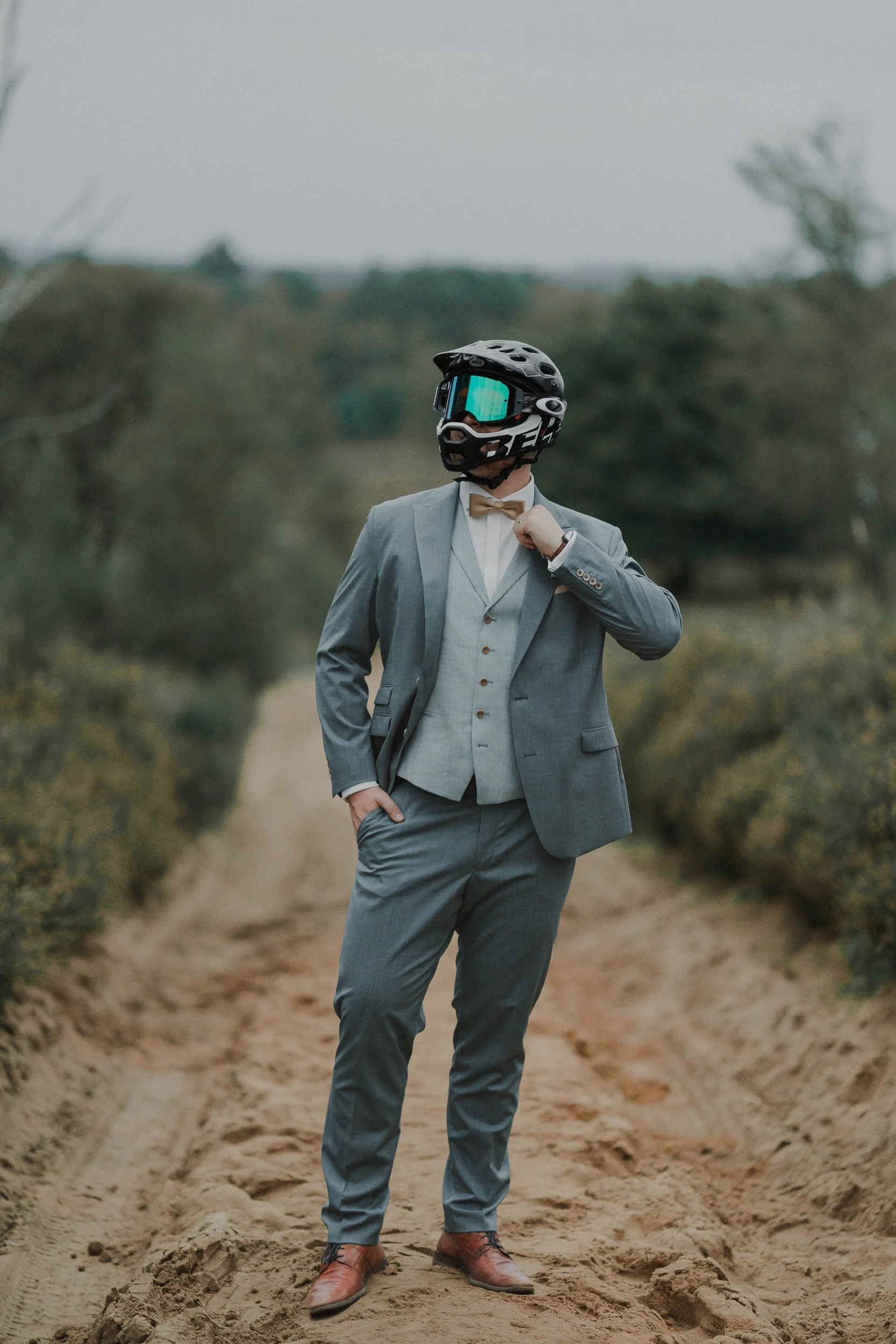 Mann in Anzug mit Fahrradhelm und Skibrille, steht auf einem sandigen Weg, auf einem Feld mit Büschen, trägt eine Fliege, hat eine Hand in der Tasche.