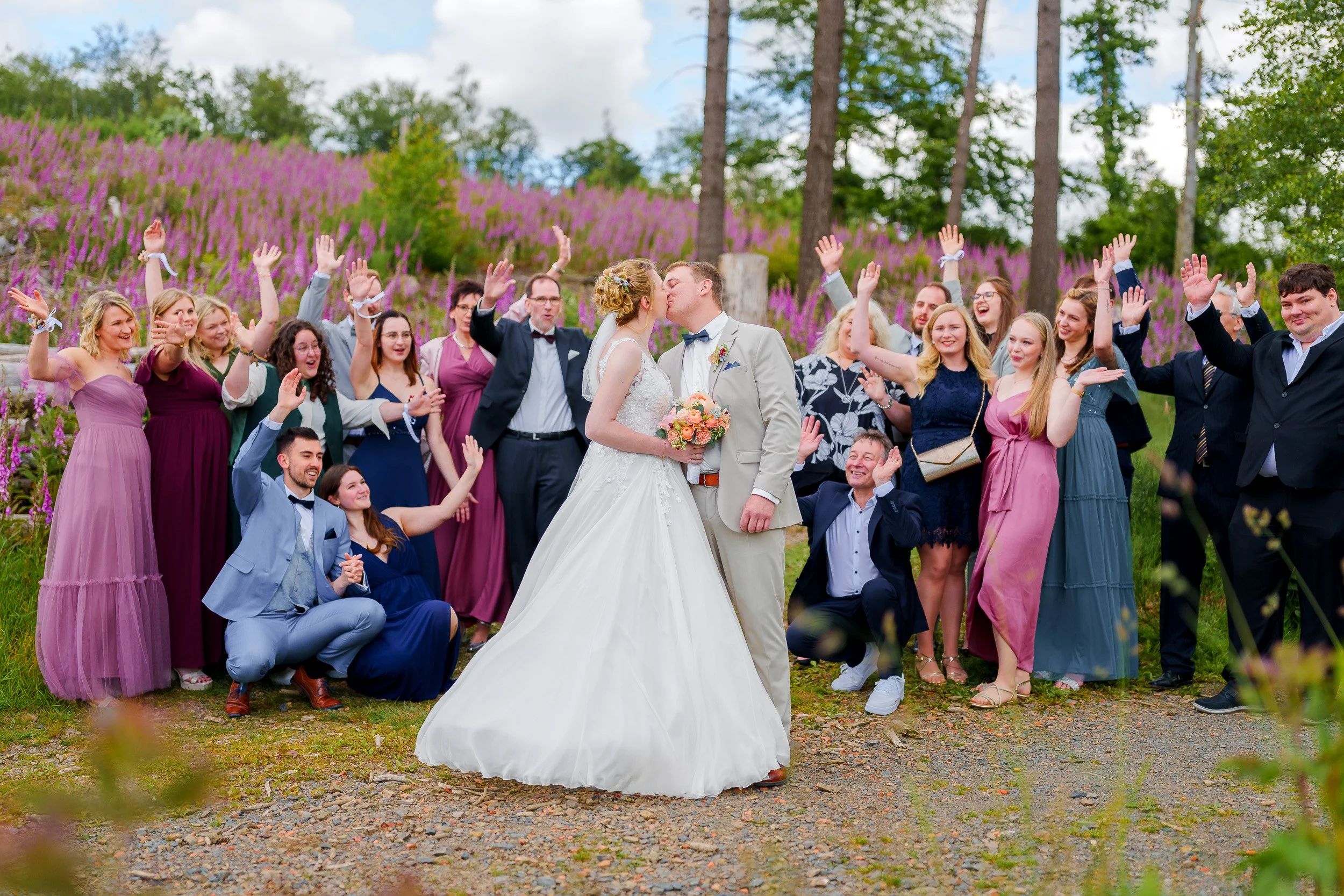 Braut und Bräutigam küssen sich während der Hochzeitsfeier im Freien, umgeben von Freunden und Familie in bunten Kleidern, mit blühenden Pflanzen im Hintergrund.