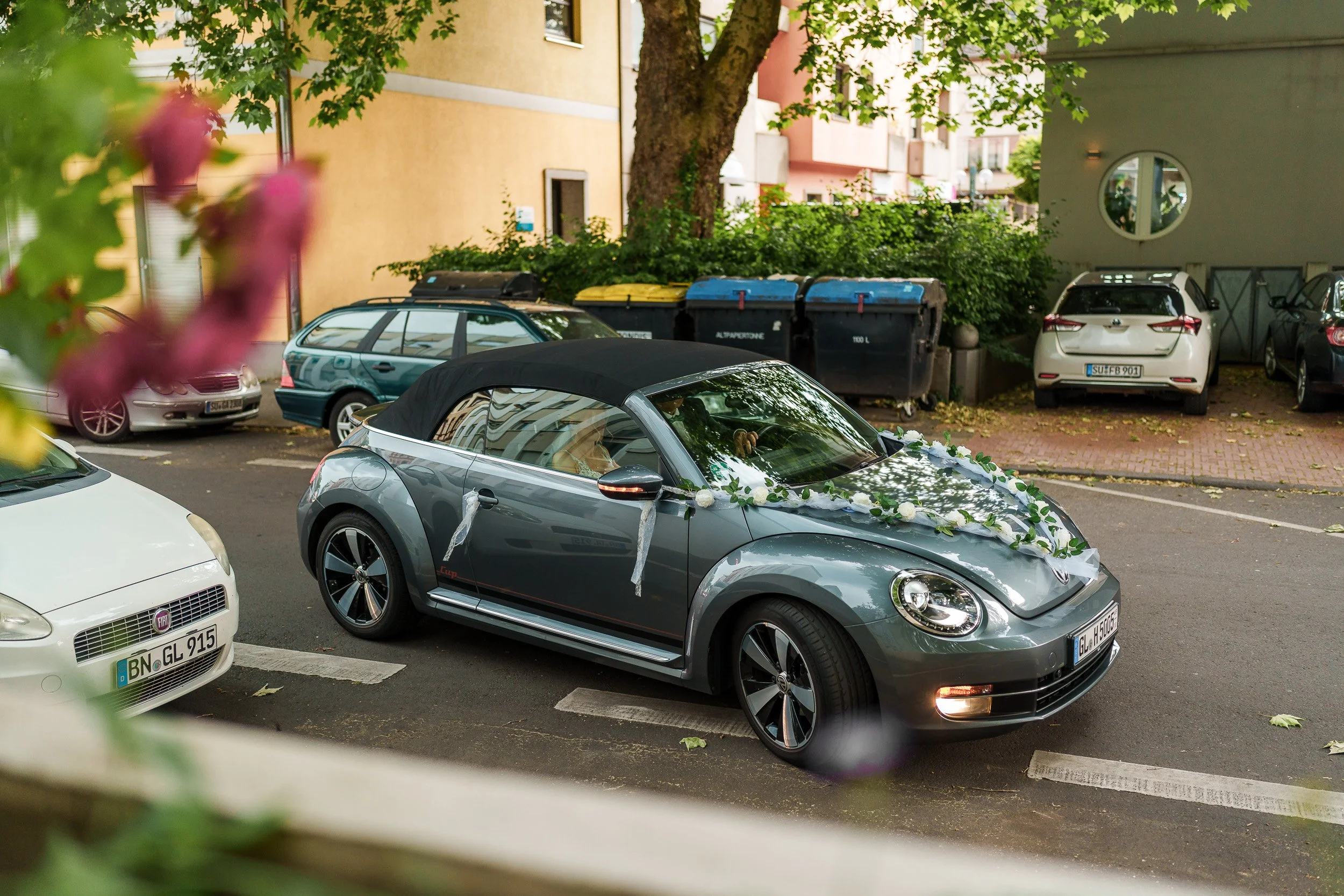 Ein grauer Volkswagen Käfer mit Hochzeitsdekoration auf der Motorhaube, parkend in einer Parkbucht, umgeben von anderen Fahrzeugen. Im Hintergrund sehen wir Mülltonnen und ein Mehrfamilienhaus.