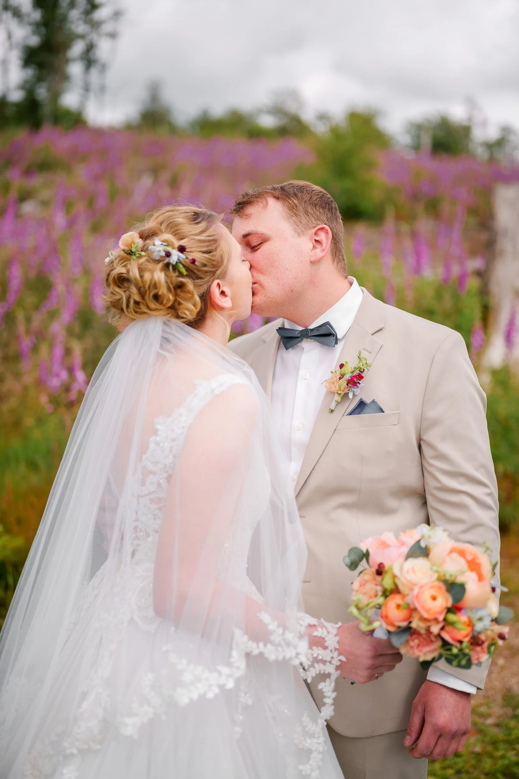 Ein Brautpaar bei einer Hochzeitszeremonie, sie küssen sich. Im Hintergrund sind blühende lila und grüne Bäume.