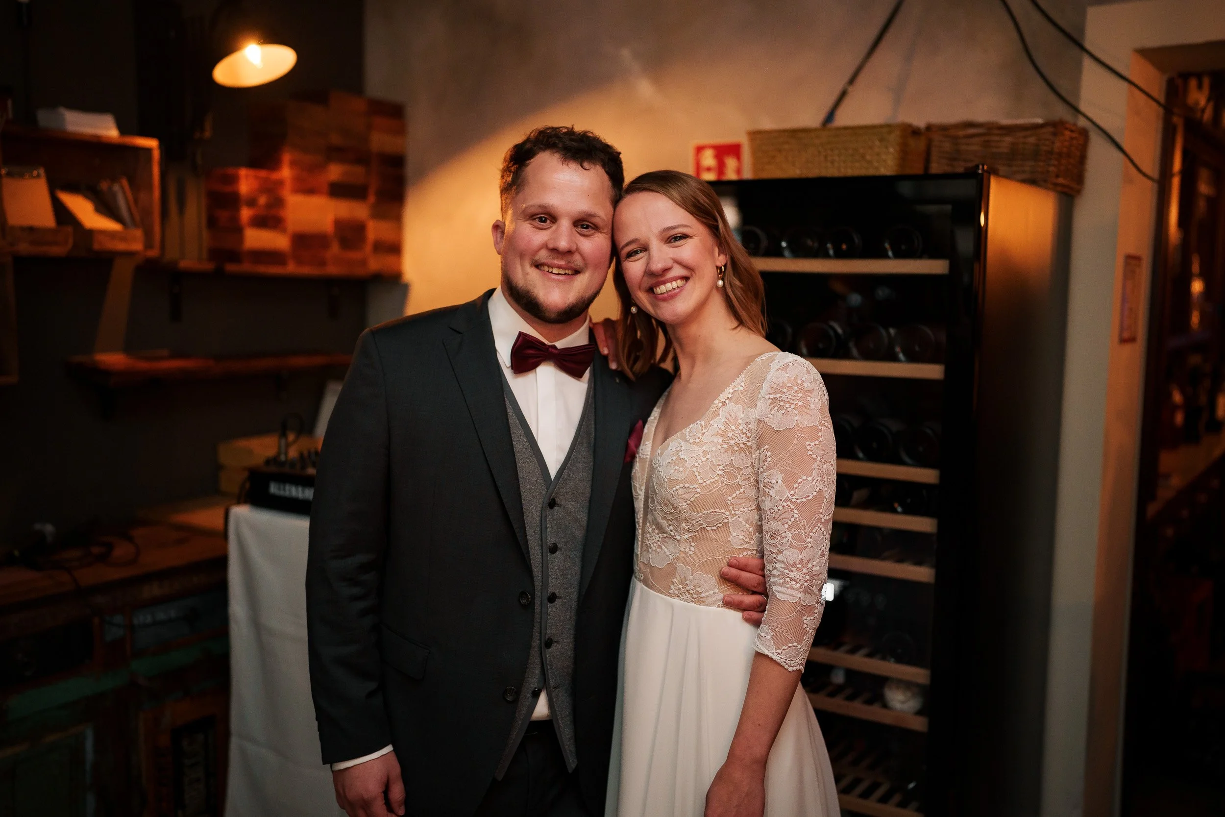 Ein glückliches Brautpaar bei einer Hochzeit, lachend, in einem festlichen Ambiente, die Frau trägt ein weißes Brautkleid mit Spitze, der Mann einen dunklen Anzug mit Fliege.