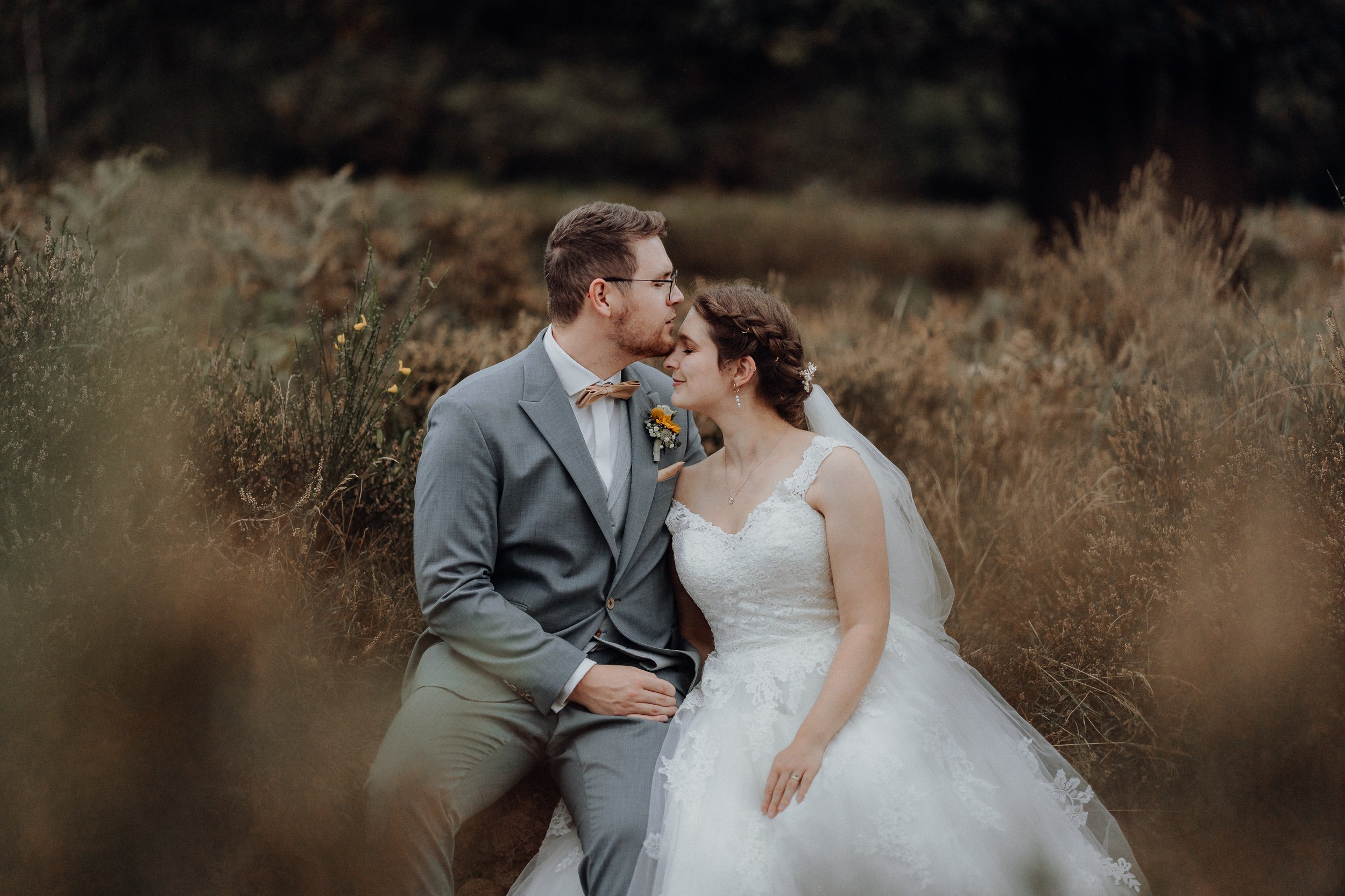 Ein Brautpaar sitzt in einer natürlichen Umgebung, sie trägt ein weißes Hochzeitskleid, er trägt einen grauen Anzug, sie kuscheln liebevoll.
