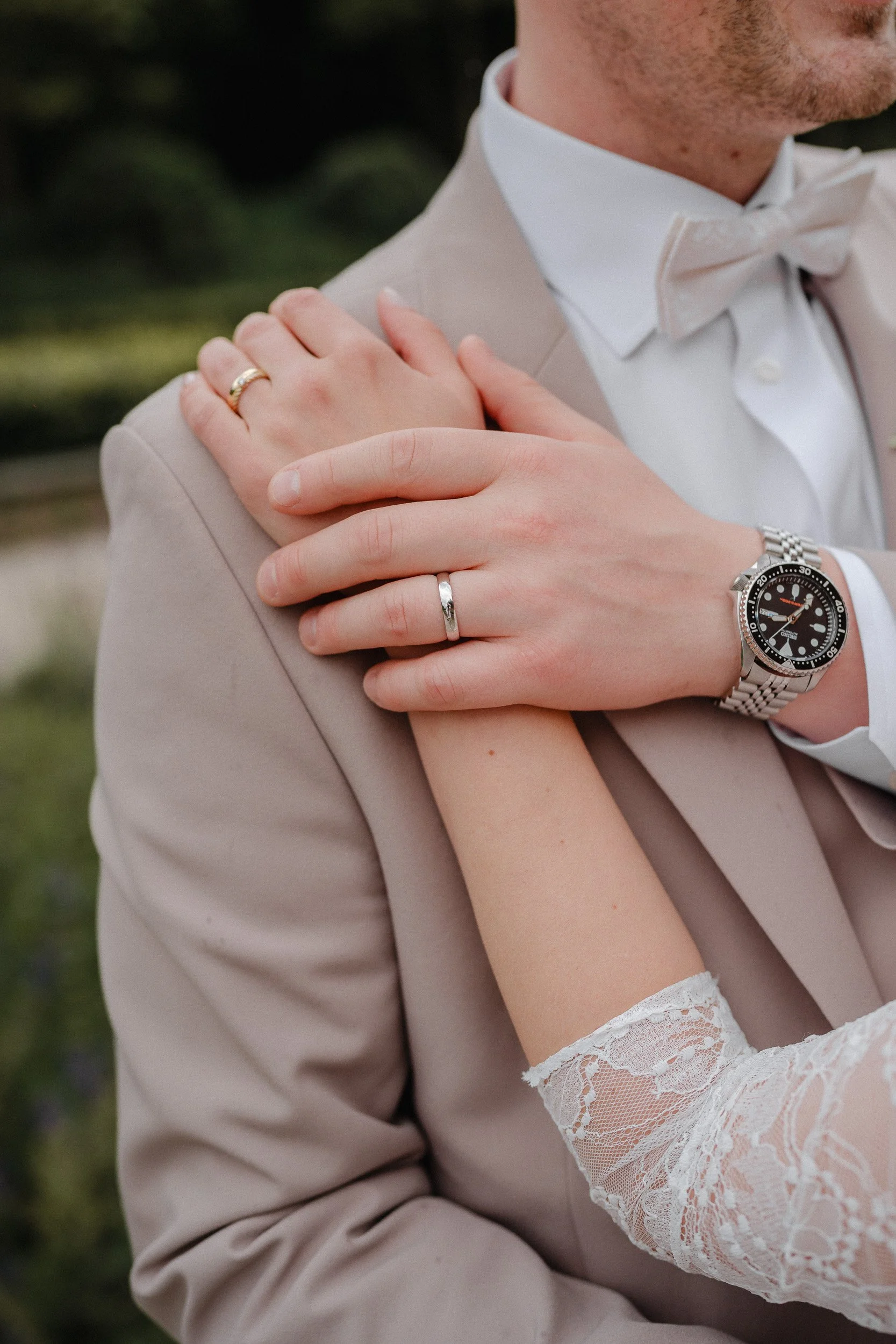 Nahaufnahme eines Paares bei einer Hochzeit, zeigt die Hände, beide tragen Eheringe. Der Mann trägt eine Armbanduhr, das Mädchen trägt ein Spitzenkleid. Hintergrund unscharf.