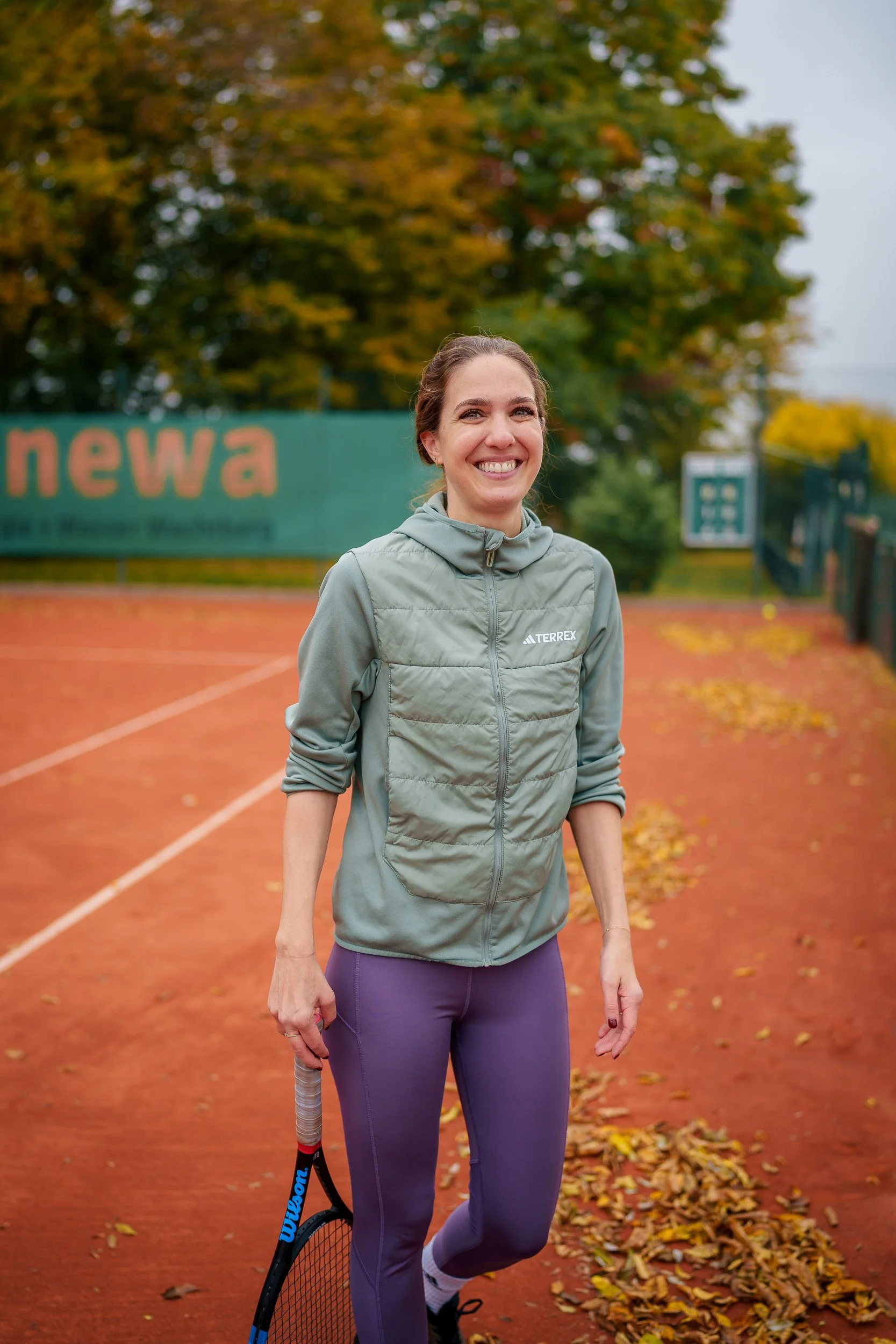 Junge Frau auf einem Tennisplatz im Herbst, trägt einen grünen Sportsweatjacke und lila Leggings, hält einen Tennisschläger, lächelt in die Kamera, bunte Blätter auf dem Boden, Bäume mit Herbstlaub im Hintergrund.
