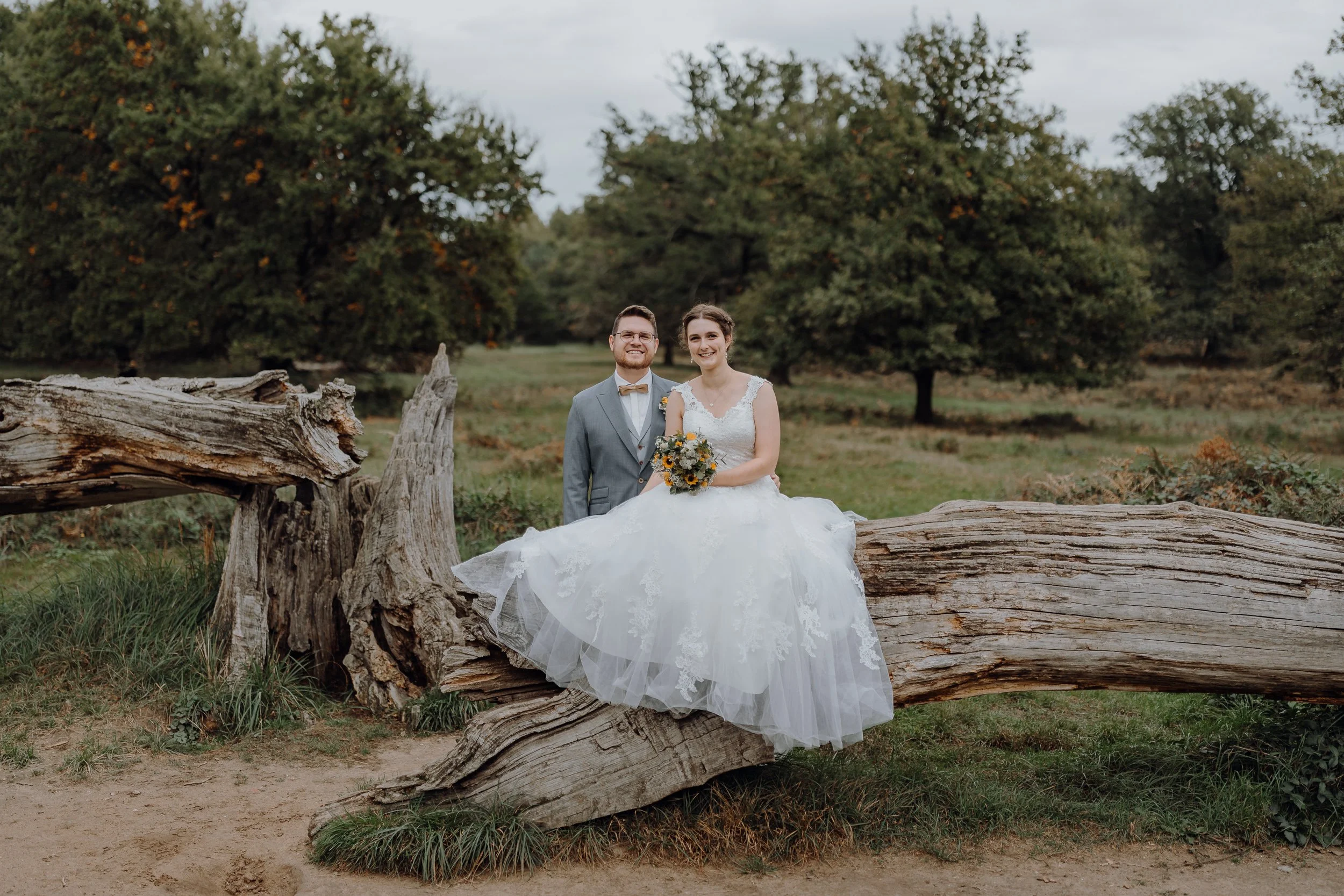 Braut und Bräutigam sitzen auf einem großen umgestürzten Baum in einem Park, umgeben von Grün und Bäumen, an einem bewölkten Tag, bei einer Hochzeit.