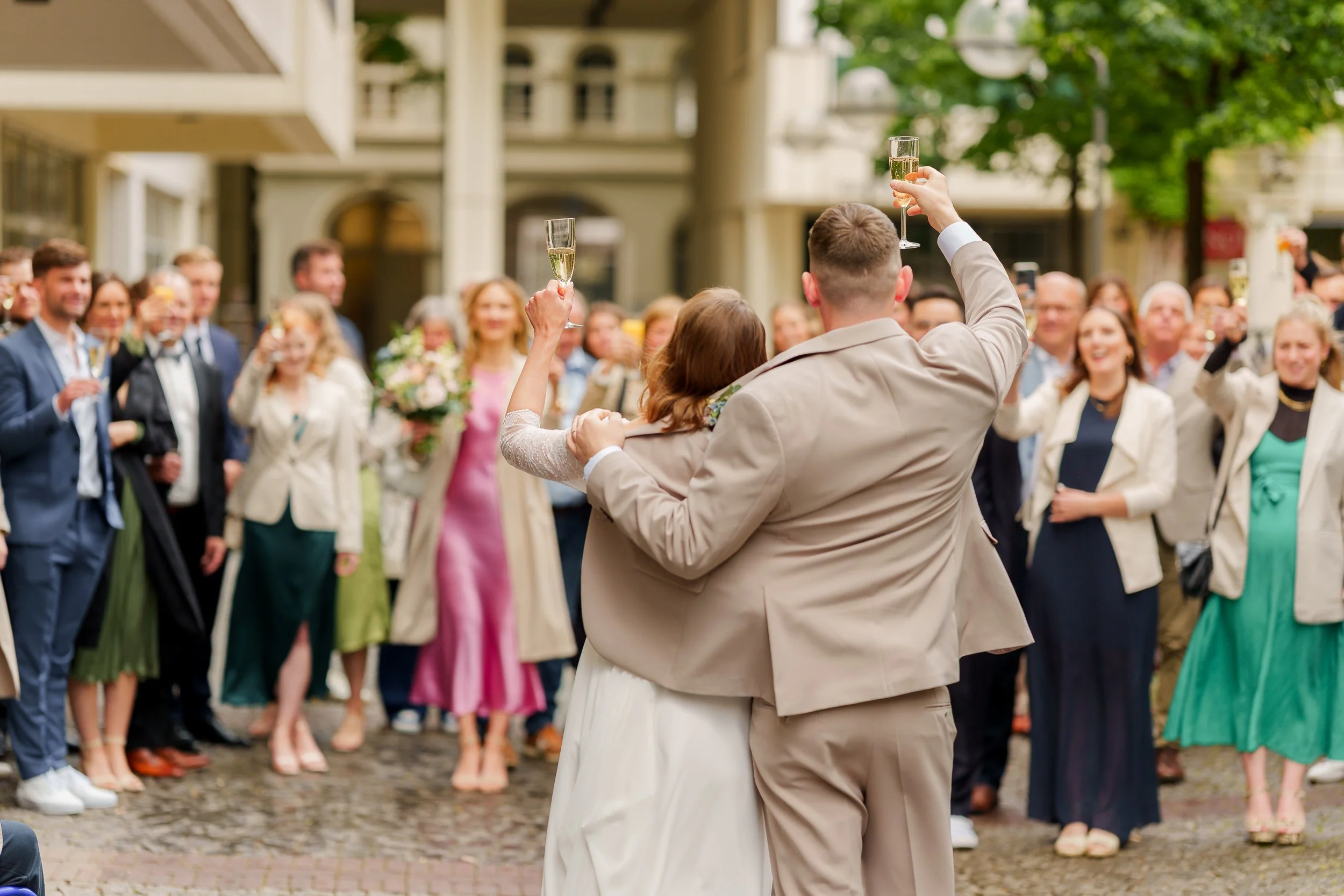 Ein frisch verheiratetes Brautpaar, das sich umarmt und mit Sekt anstößt, während eine große Gruppe von Freunden und Familie zuschaut und feiert.