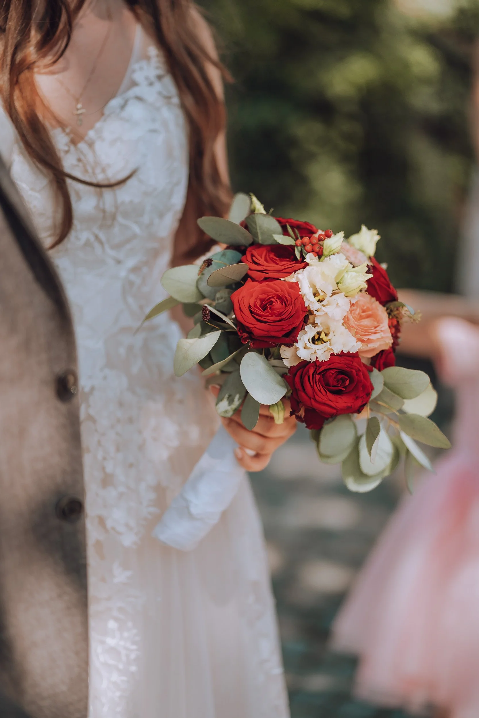 Eine Braut hält einen Blumenstrauß aus roten und rosa Rosen, weißen Orchideen und grünem Eukalyptus bei einer Hochzeit.