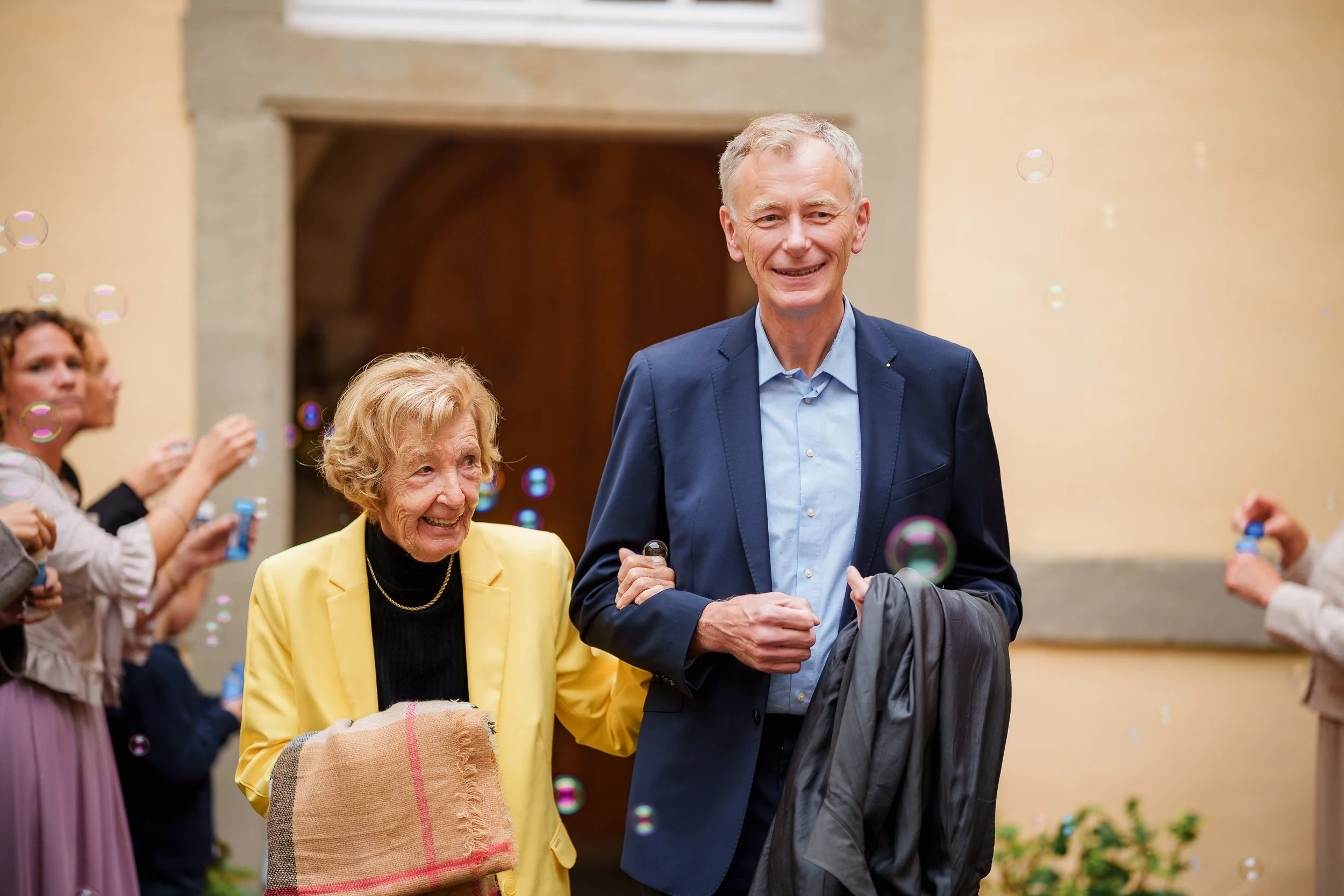 Ein älterer Mann und eine ältere Frau feiern, umgeben von Freunden, mit Seifenblasen im Hintergrund.
