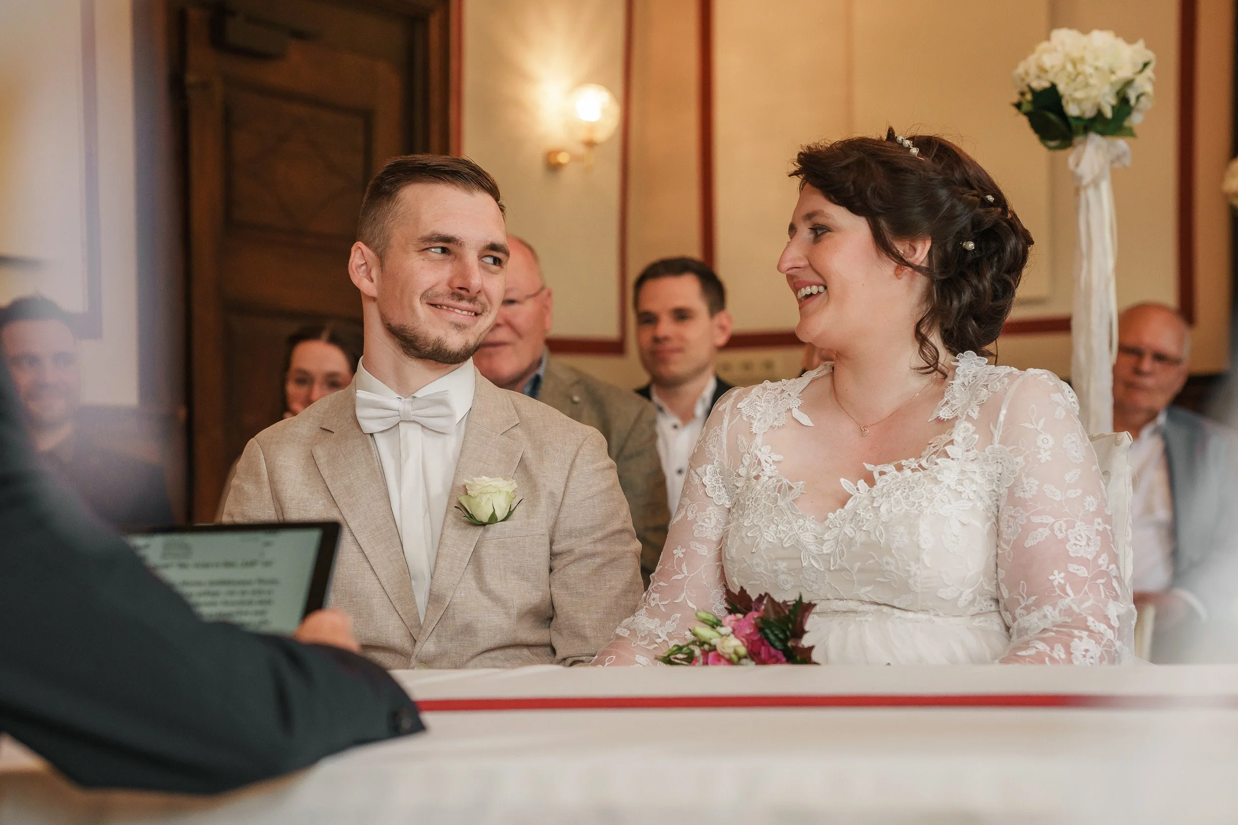 Ein Brautpaar bei ihrer Trauung, beide lächeln sich an und sitzen gegenüber. Im Hintergrund sitzen weitere Gäste im festlichen Raum.