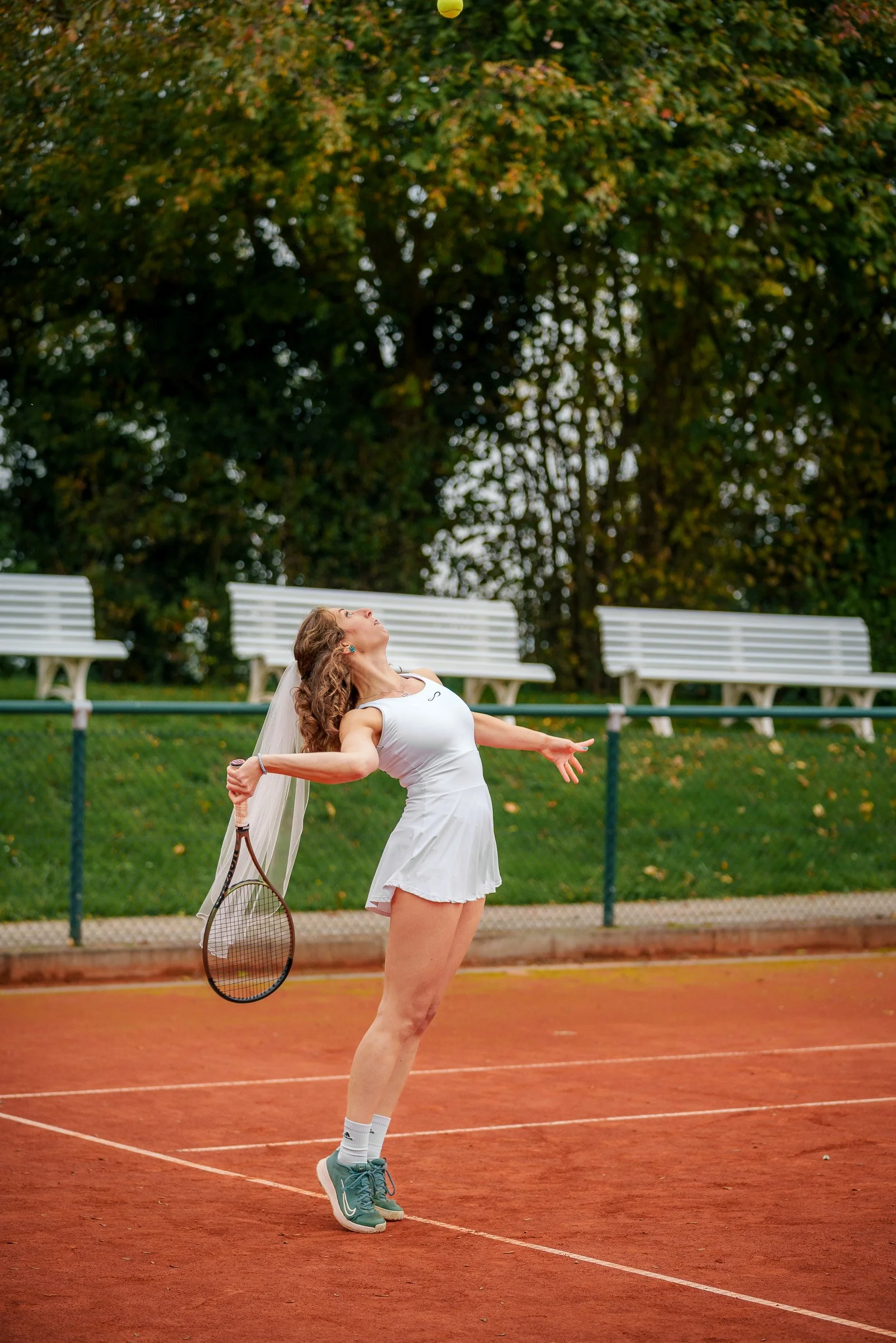 Frau auf einem Tennisplatz beim Aufschlag, trägt weiße Sportkleidung und Schuhe, hält einen Tennisball und einen Tennisschläger, mit Bänken und Bäumen im Hintergrund.