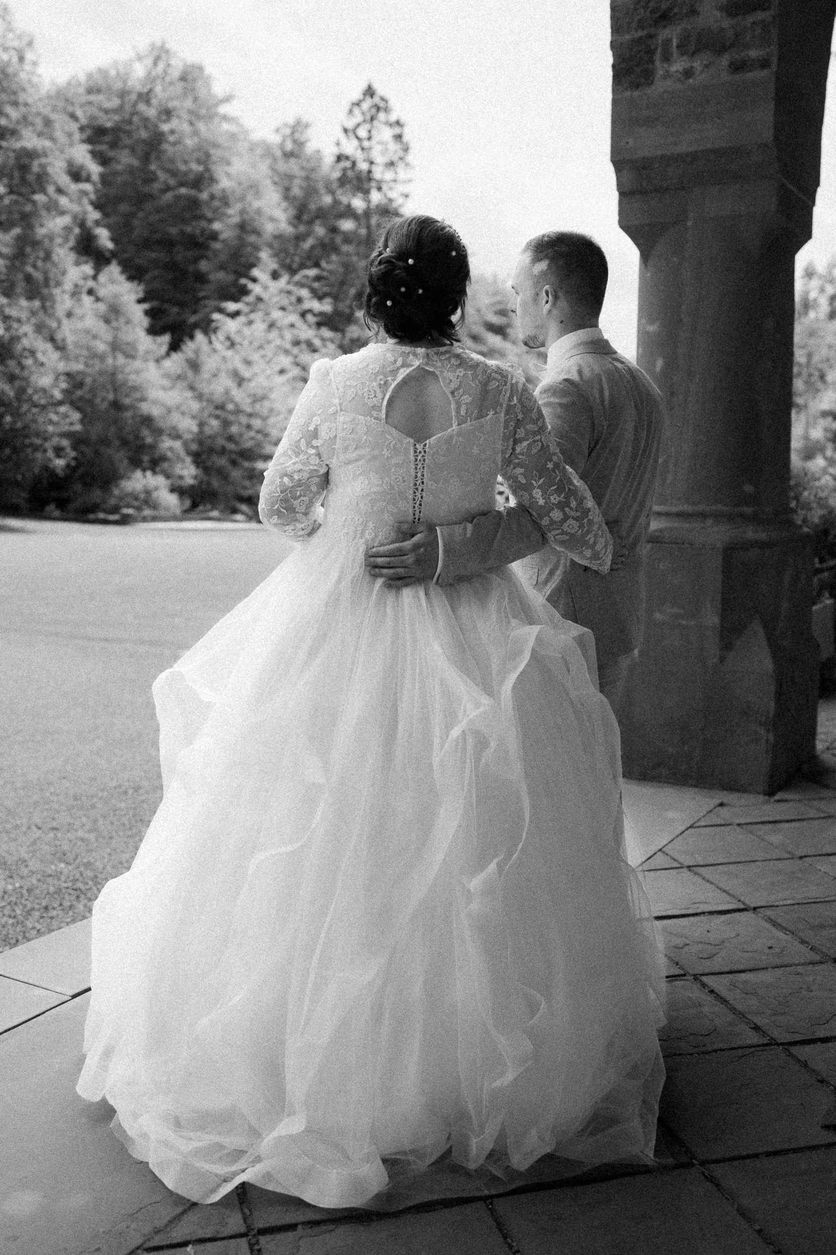 Braut und Bräutigam stehen zusammen im Freien vor einer Kirche, die Frau trägt ein langes, beiges Brautkleid mit Spitzenärmeln, der Mann trägt einen hellen Anzug. Im Hintergrund sind Bäume zu sehen.