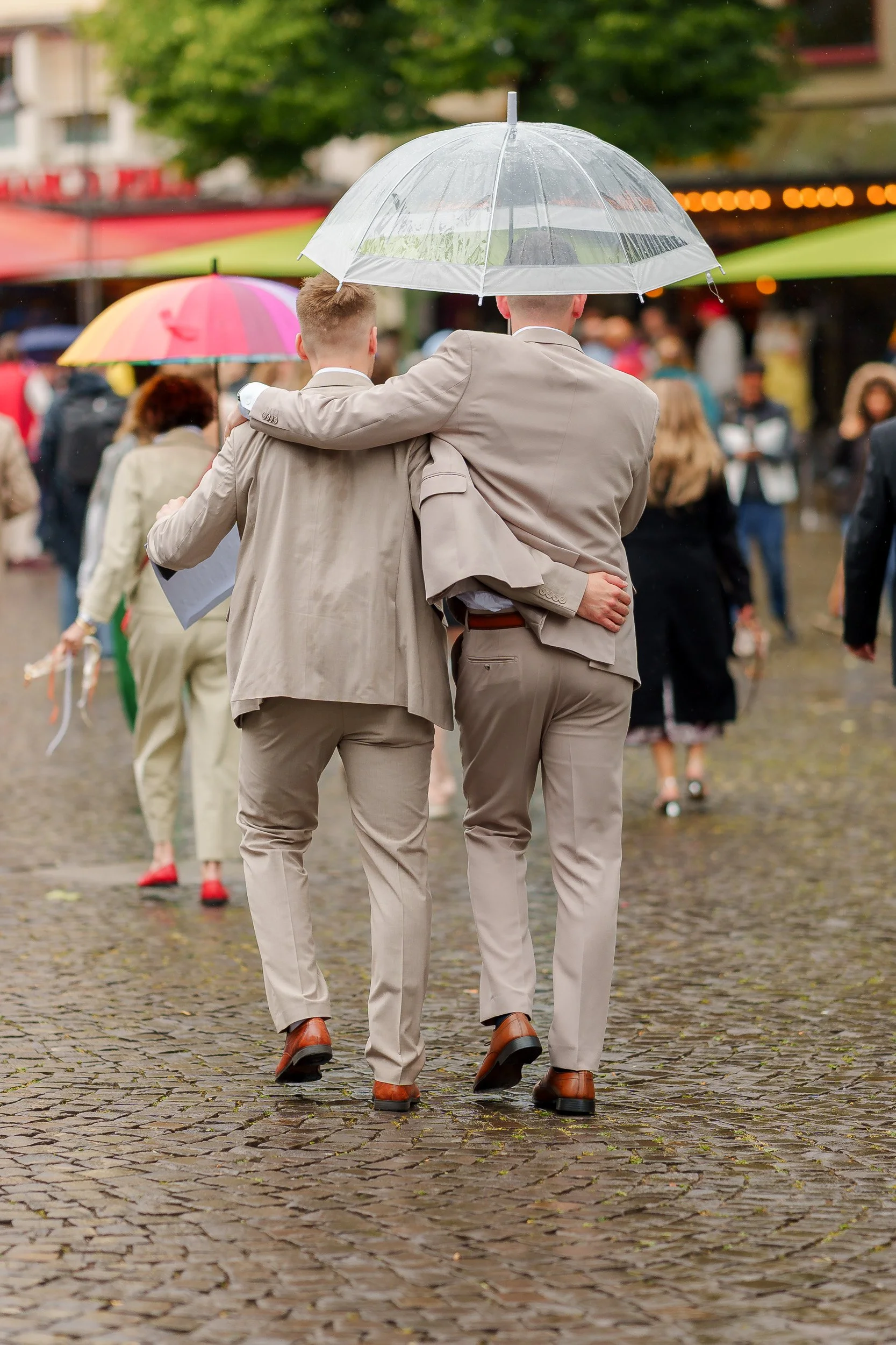Zwei Männer in Anzügen sitzen nebeneinander und haben ihre Arme um die Schulter des jeweils anderen, während sie bei Regen unter einem transparenten Regenschirm auf einer belebten, nassen Straße gehen.