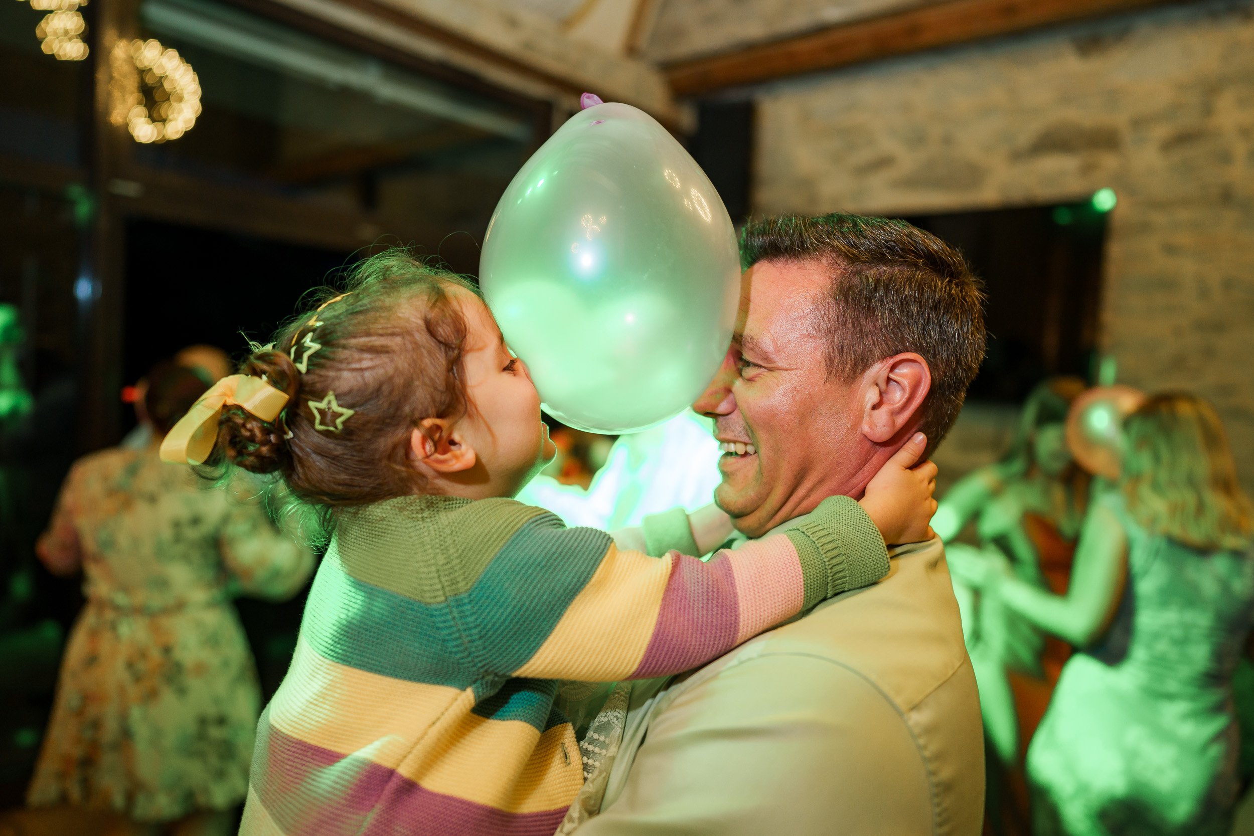 Ein lachender Mann hält ein lachendes kleines Mädchen, das ein Ballon berührt, bei einer Feier im Innenraum, umgeben von anderen feiernden Menschen.
