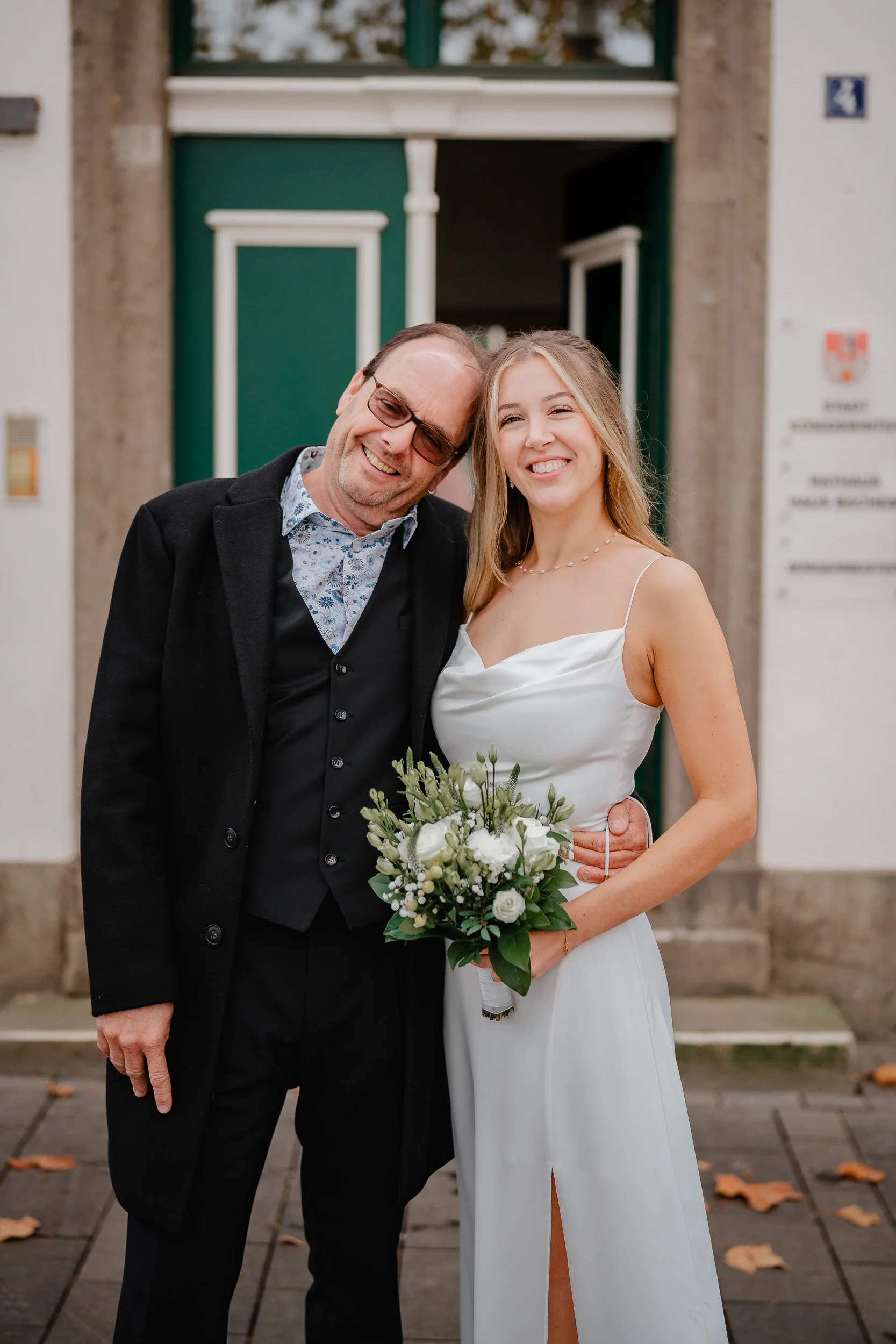 Ein Mann und eine junge Frau stehen vor einem Gebäude, lächeln und umarmen sich. Die Frau hält einen Blumenstrauß. Es ist vermutlich eine Hochzeit.