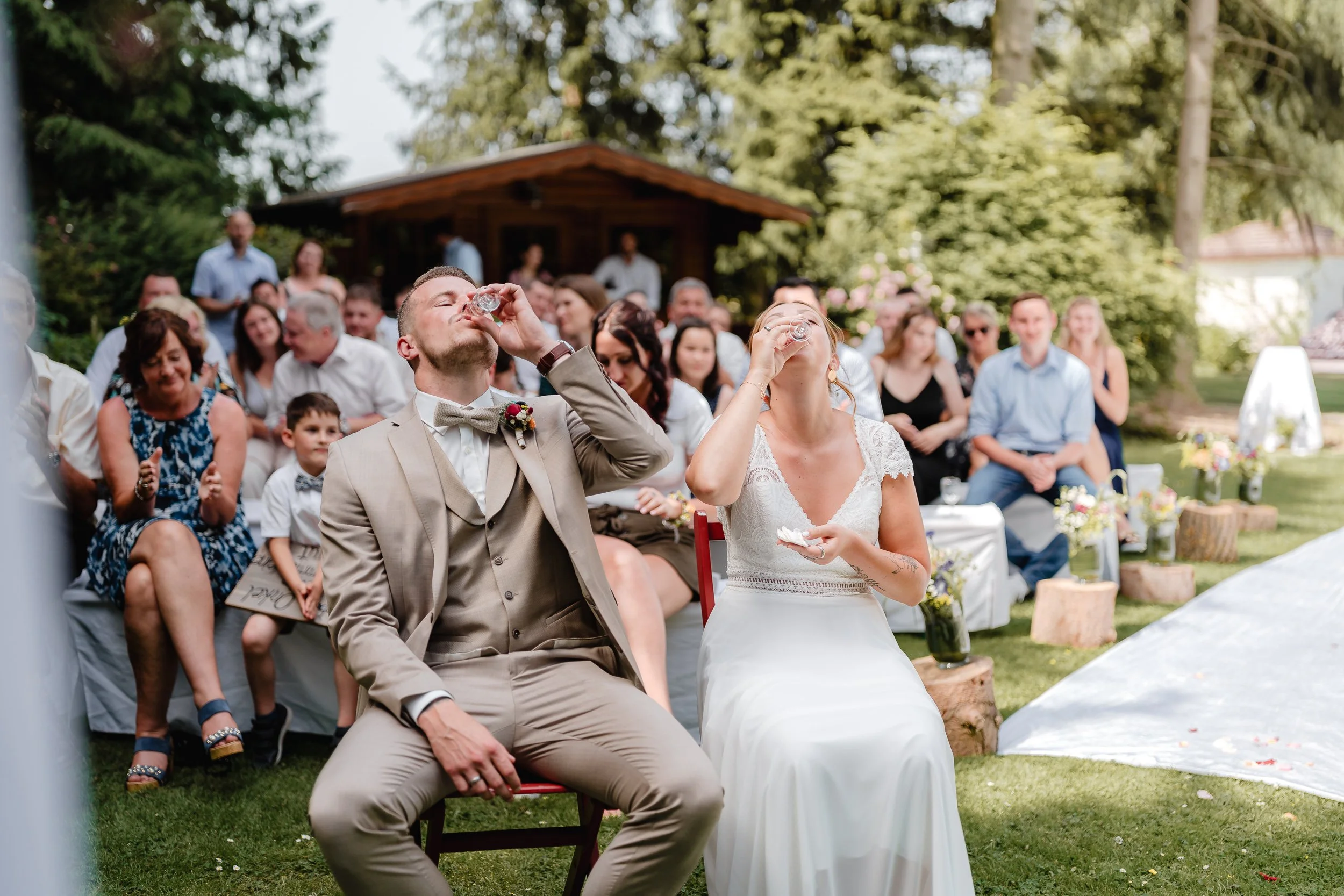 Braut und Bräutigam beim Sektempfang während der Hochzeit, sie trinken an ihren Sitzplätzen im Garten, im Hintergrund sitzt das Hochzeitsgäste, die zuschauen.