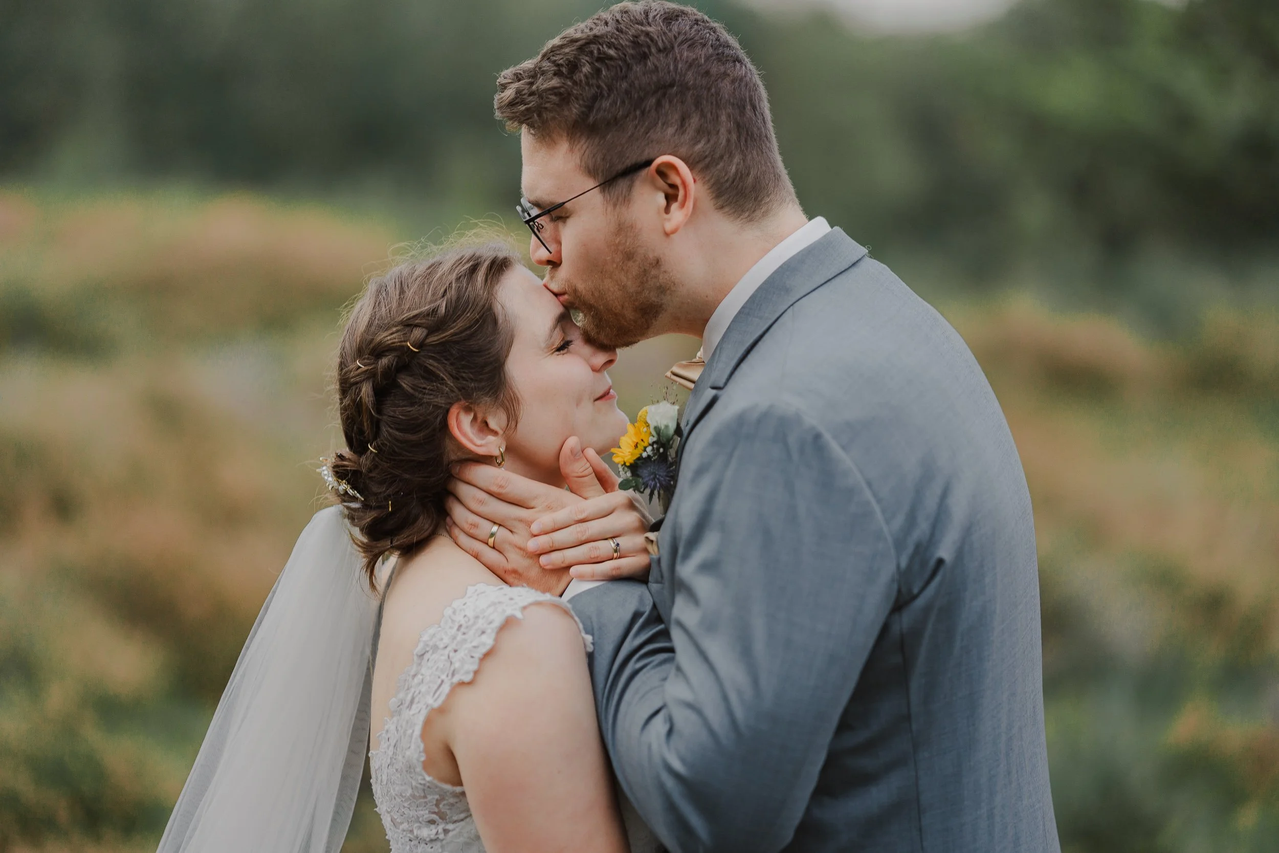 Ein Brautpaar beim Kuss, die Frau trägt ein Brautkleid mit Spitzenärmel, der Mann trägt einen grauen Anzug mit Fliege, im Hintergrund eine grüne Landschaft.