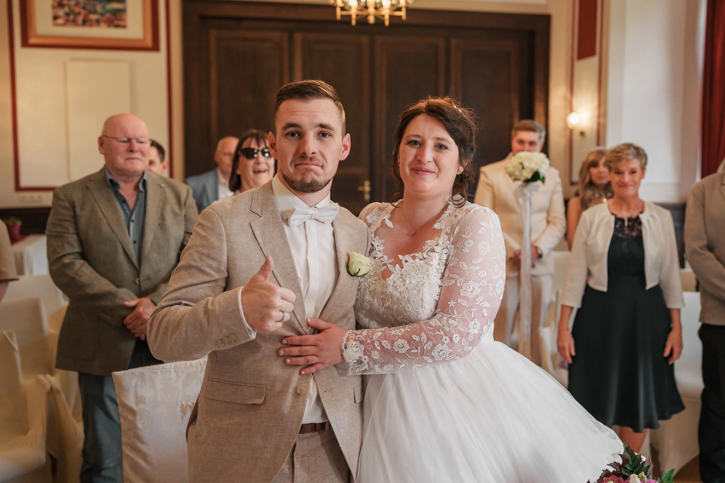 Ein Brautpaar steht in einem Saal, umgeben von Gästen. Der Bräutigam trägt einen beige Anzug mit Fliege, die Braut ein weißes Brautkleid mit Spitze. Die Gäste hinter ihnen lächeln und applaudieren.