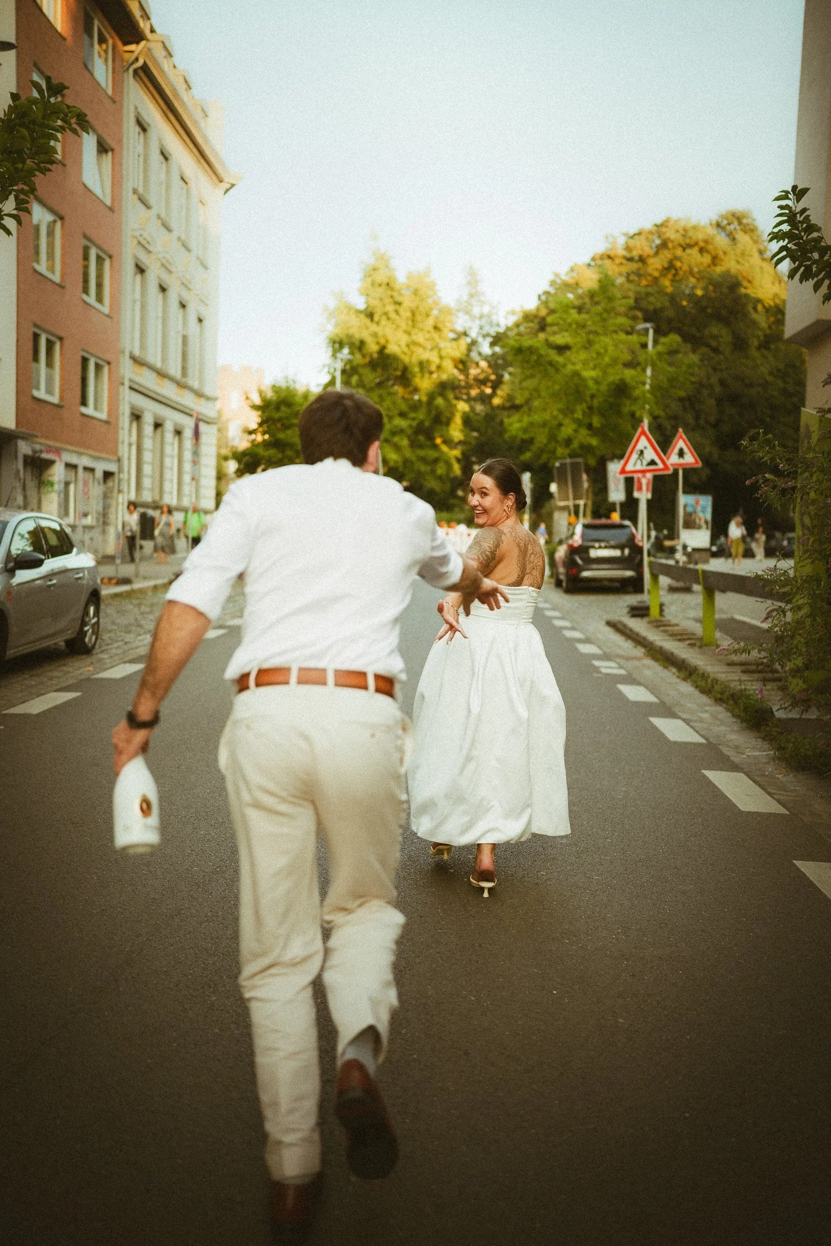 Ein Mann läuft lachend hinter einer Frau auf der Straße, die in einem weißen Kleid auf Rollschuhen fährt, bei Sonnenuntergang, umgeben von Stadtgebäuden und Bäumen.