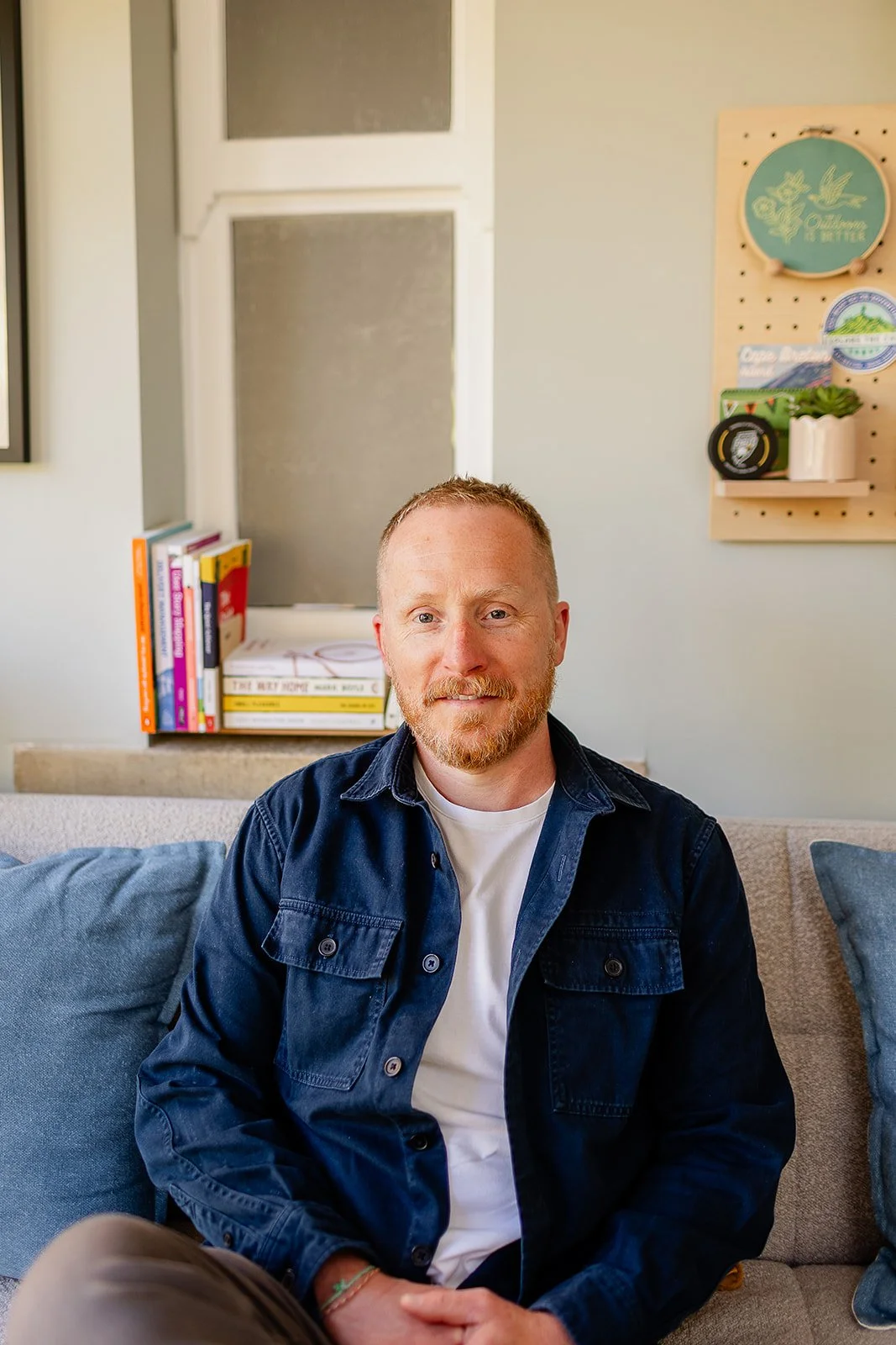 A man sitting on a sofa smiling at the camera. There's a book shelf behind him with books about delivery management, content design and research