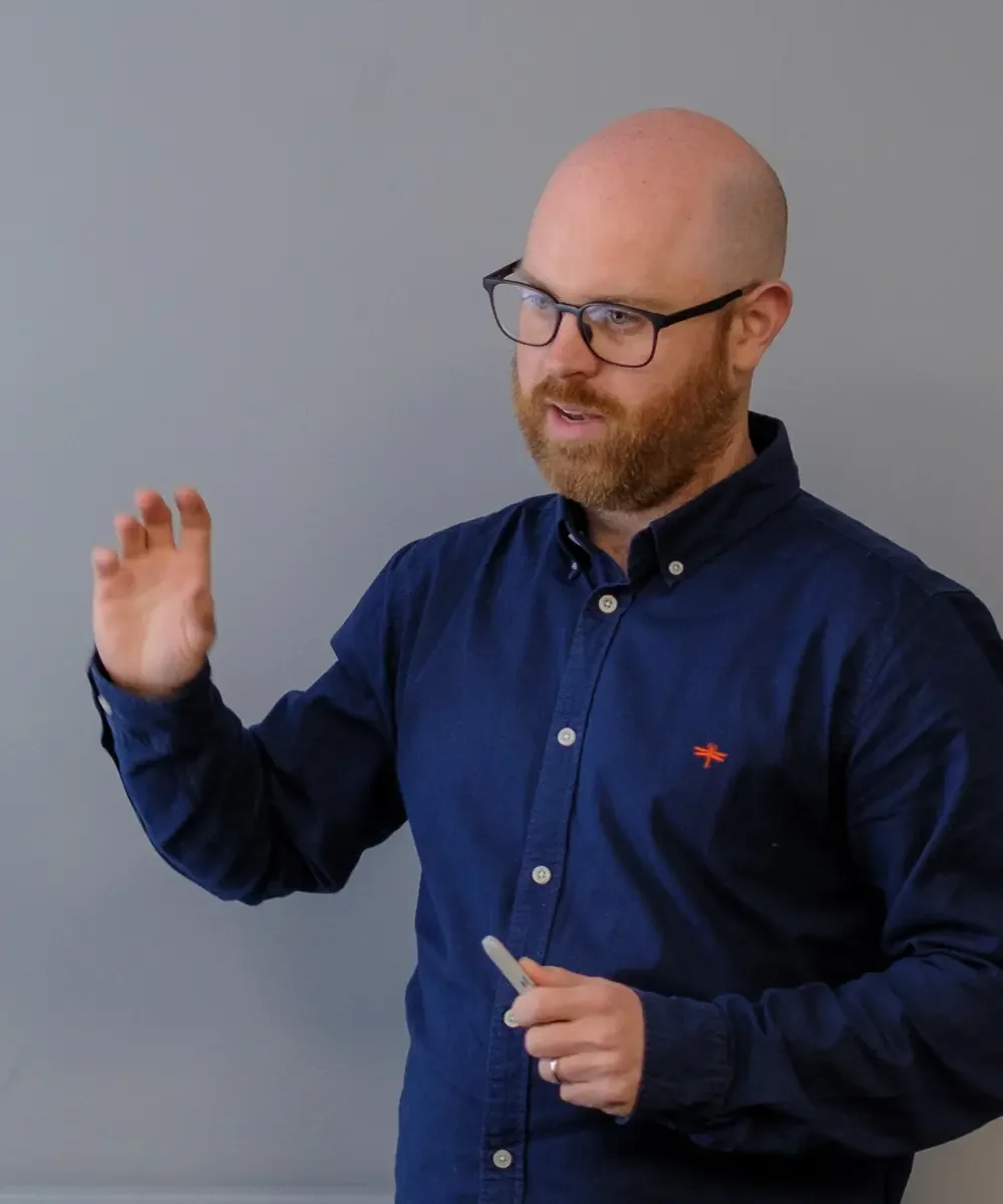 A bald man with glasses, wearing a navy blue button-up shirt, is talking and gesturing with his right hand, holding a marker in his left hand, standing against a plain gray wall.