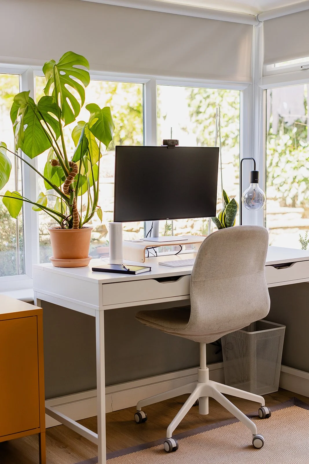 Home office desk with a computer monitor, potted plants, notebook, and a hanging light bulb, with a beige office chair in front, situated near large windows with greenery outside.
