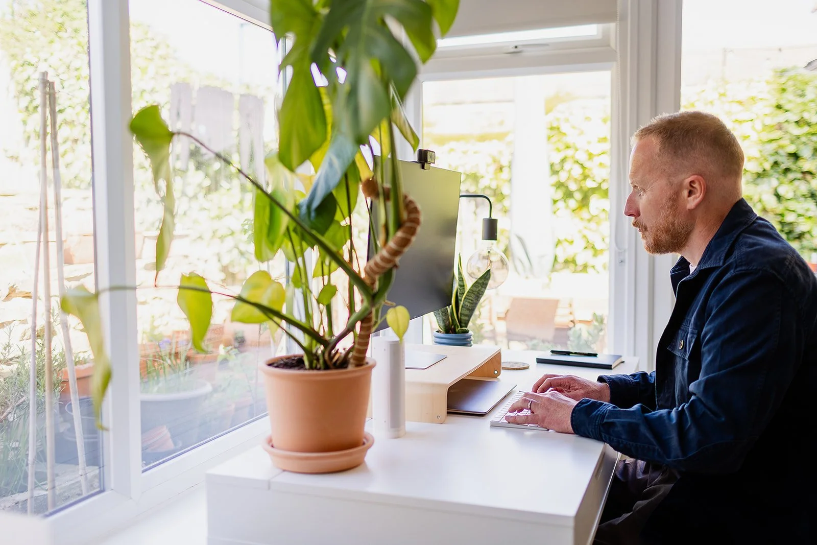 Man working at a white desk near large windows with potted plants and greenery outside.