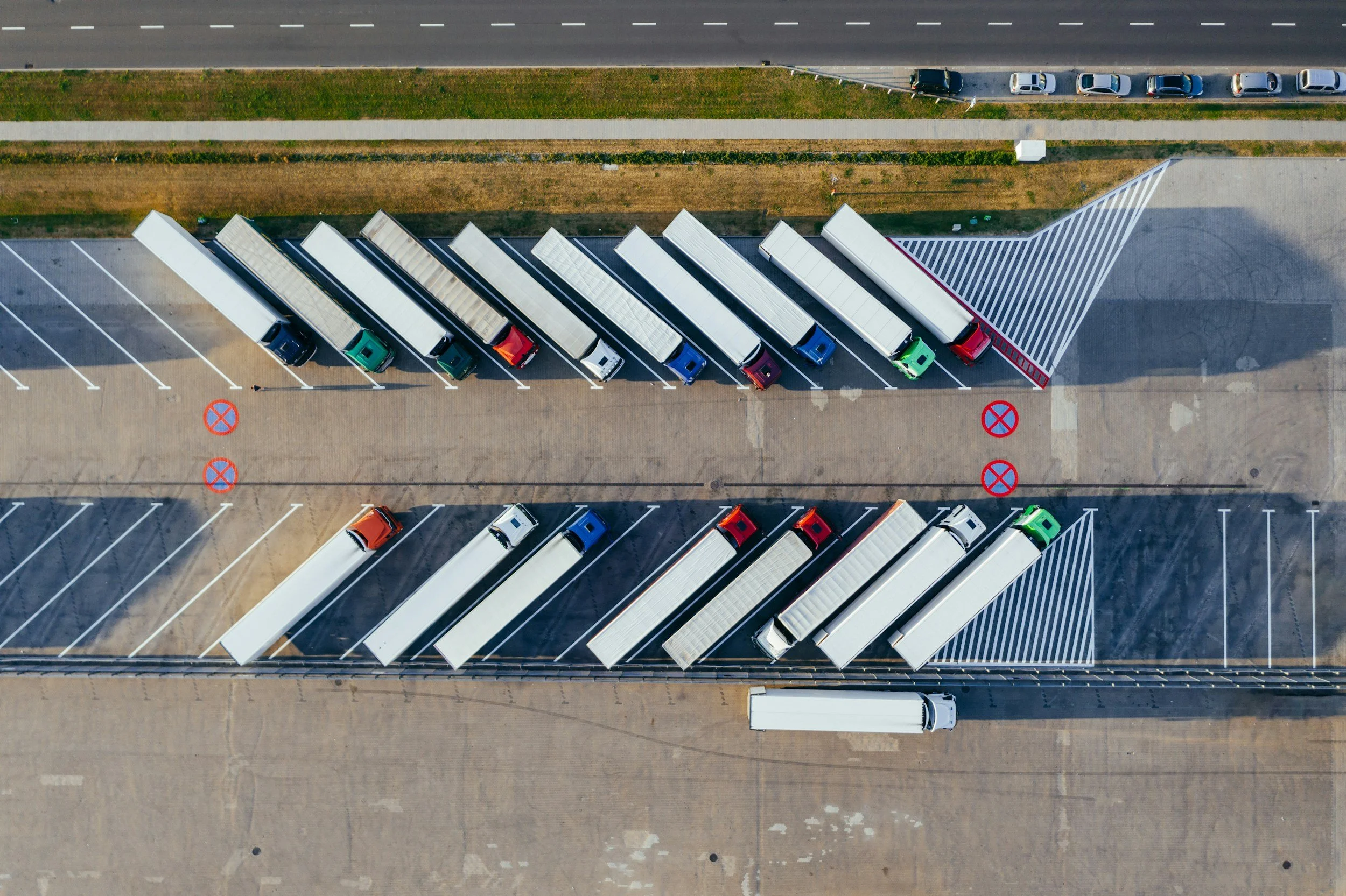 An aerial view of a parking lot with multiple rows of trucks and cars, adjacent to a green and brown grassy area separated by a sidewalk. The parking lot has marked spaces and no visible people.