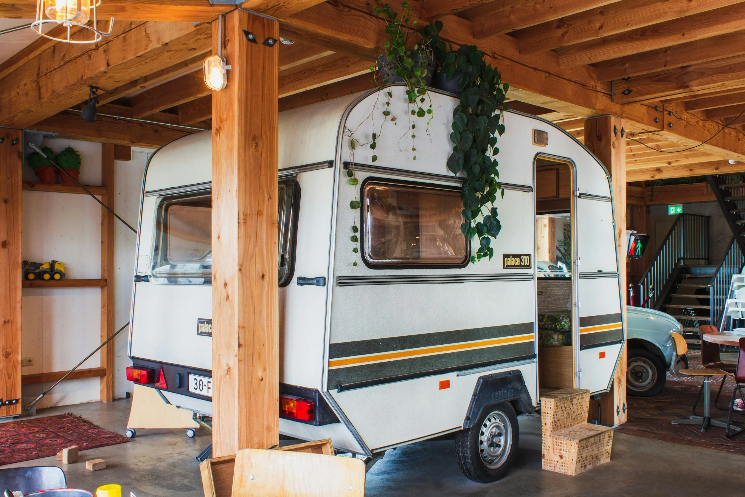 Interior of a wooden structure with a vintage camper trailer, potted plants, and various decorative items. The ceiling has exposed wooden beams and lighting fixtures. There are stairs and a crosswalk sign visible in the background.