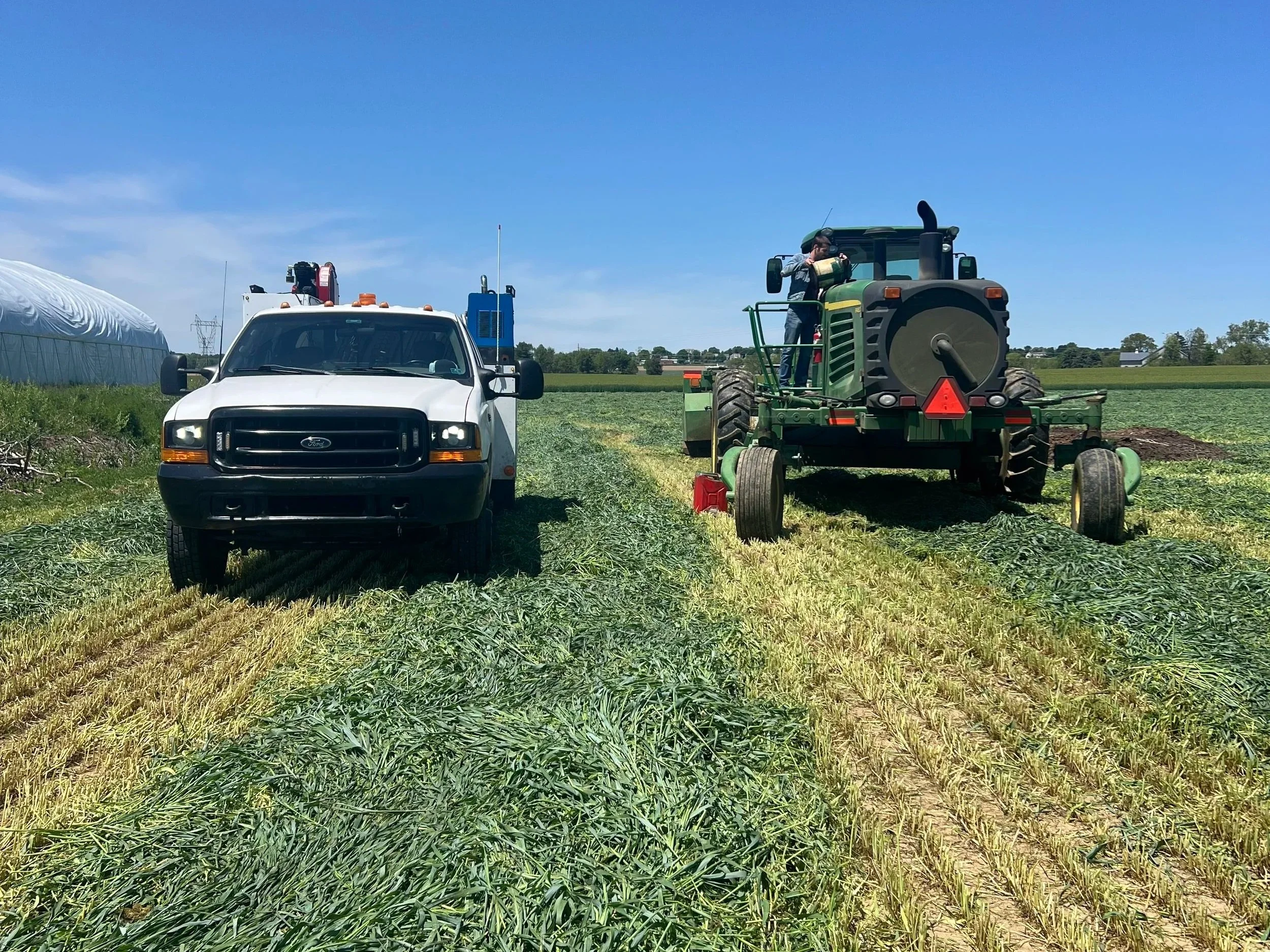 A person operating a green tractor in a field next to a white pickup truck with machinery mounted on it, under a clear blue sky.