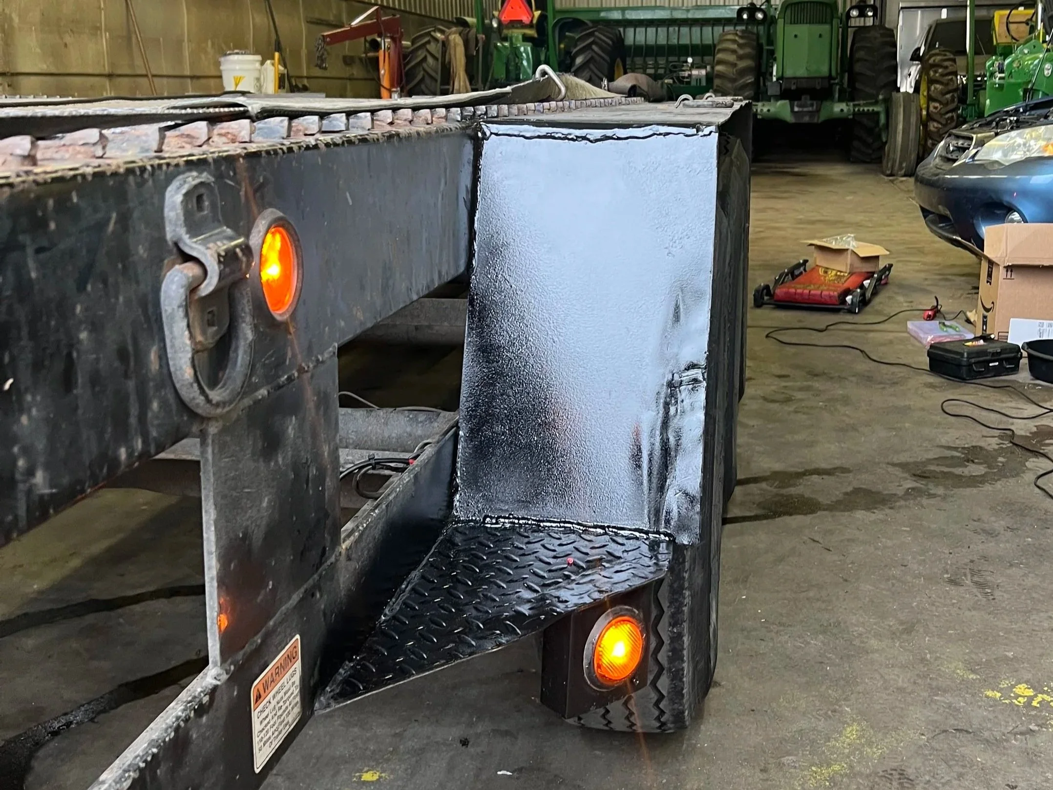 A flatbed trailer with a black surface and orange lights is parked inside a storage or workshop area, with various tools, machinery, and tractors seen in the background.
