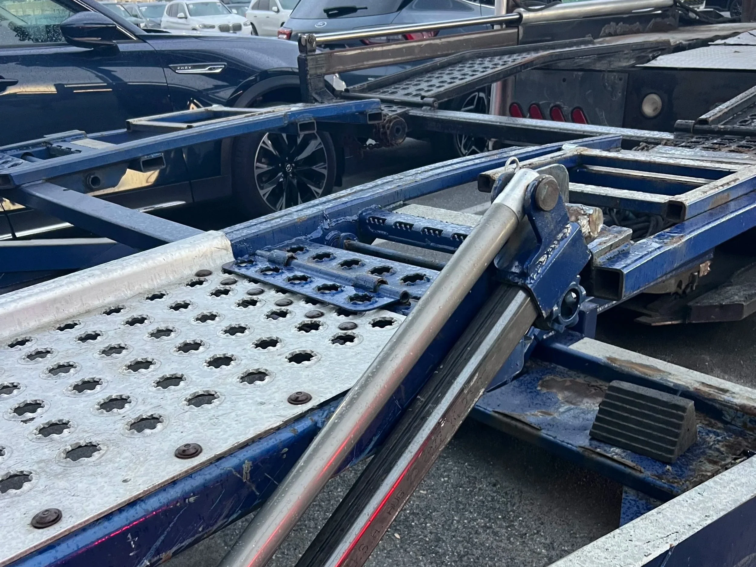 Close-up of metal safety rails and steps on a flatbed trailer, with a blue car and other vehicles in a parking lot in the background.