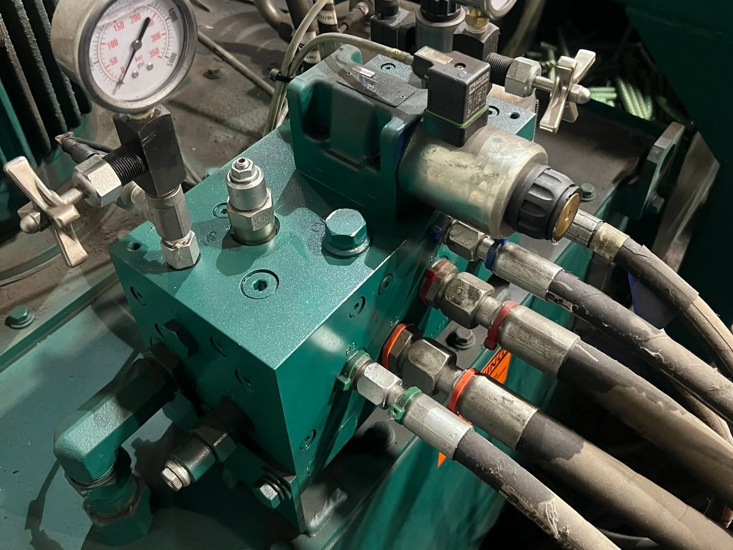 Close-up of a hydraulic or industrial machine component featuring a pressure gauge, various metal pipes, fittings, and adjustment valves on a teal metallic base.